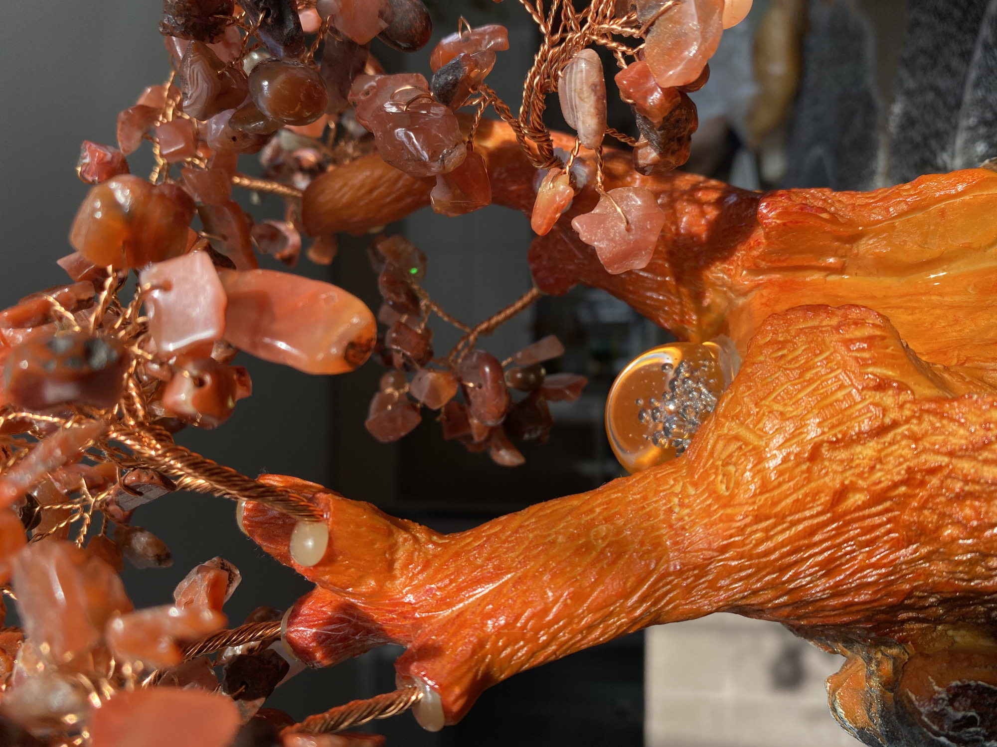 The image presents a close-up view of a decorative tree-like sculpture, showcasing its intricate details and materials. The sculpture features numerous small, polished, and translucent stones in various shades of reddish-orange and amber, resembling leaves or fruit. These stones are attached to delicate branches made of twisted copper wire. Some stones exhibit natural banding.
The main trunk and larger branches of the tree are sculpted from a solid, vibrant orange material, which appears to be resin or a similar substance, textured to mimic bark or wood grain. Small, spherical, opaque white beads are integrated into parts of this orange structure. Additionally, there are clear, circular elements embedded in the sculpture, filled with tiny, reflective silver beads that catch the light.
The lighting is bright, creating strong highlights and reflections on the stones, copper wire, and the glossy surface of the sculpted trunk, suggesting a well-illuminated indoor setting. The background is dark and indistinct, ensuring the focus remains on the sculpture. No specific activity is depicted, as it is a static object. There is no visible text. This media file was taken in Фоки, Russia.