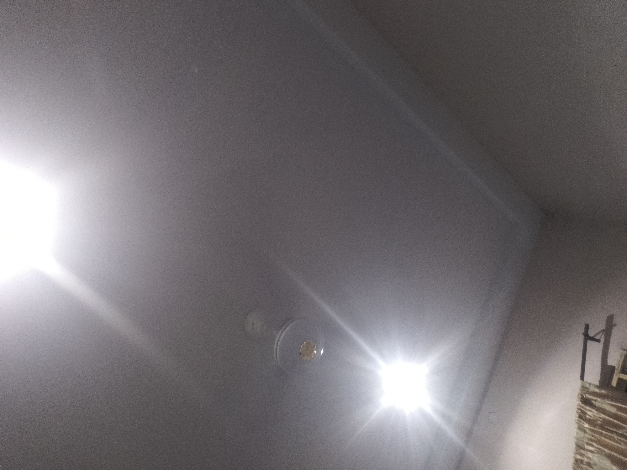 A ceiling fan with a light fixture is visible on a textured ceiling. A bright light source, likely a flash, illuminates the scene from the right, creating strong lens flares and washing out parts of the image. The ceiling appears to be a neutral color, possibly white or light gray, with subtle shadows and highlights indicating the contours of the room. To the far right, a portion of a brick structure and a dark metal object are partially visible, suggesting the presence of other elements in the environment outside the immediate focus of the ceiling. There are no people or specific details about the location that can be discerned from the image.