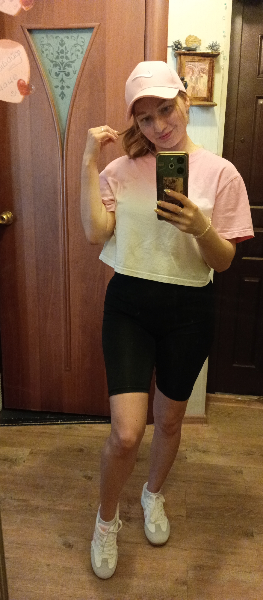 A woman is standing in what appears to be the interior of a residence. She is taking a selfie with a mobile phone. The woman is wearing a light pink baseball cap with a white Nike swoosh, a two-tone t-shirt with a pink top and a white bottom, black bike shorts, and white sneakers. She is holding a strand of her hair with her left hand. The background features two wooden doors of different shades and designs. On the wall, there is a decorative shelf holding a small framed picture and some pinecones. The floor is made of wood. There is a sticker on the door on the left.
