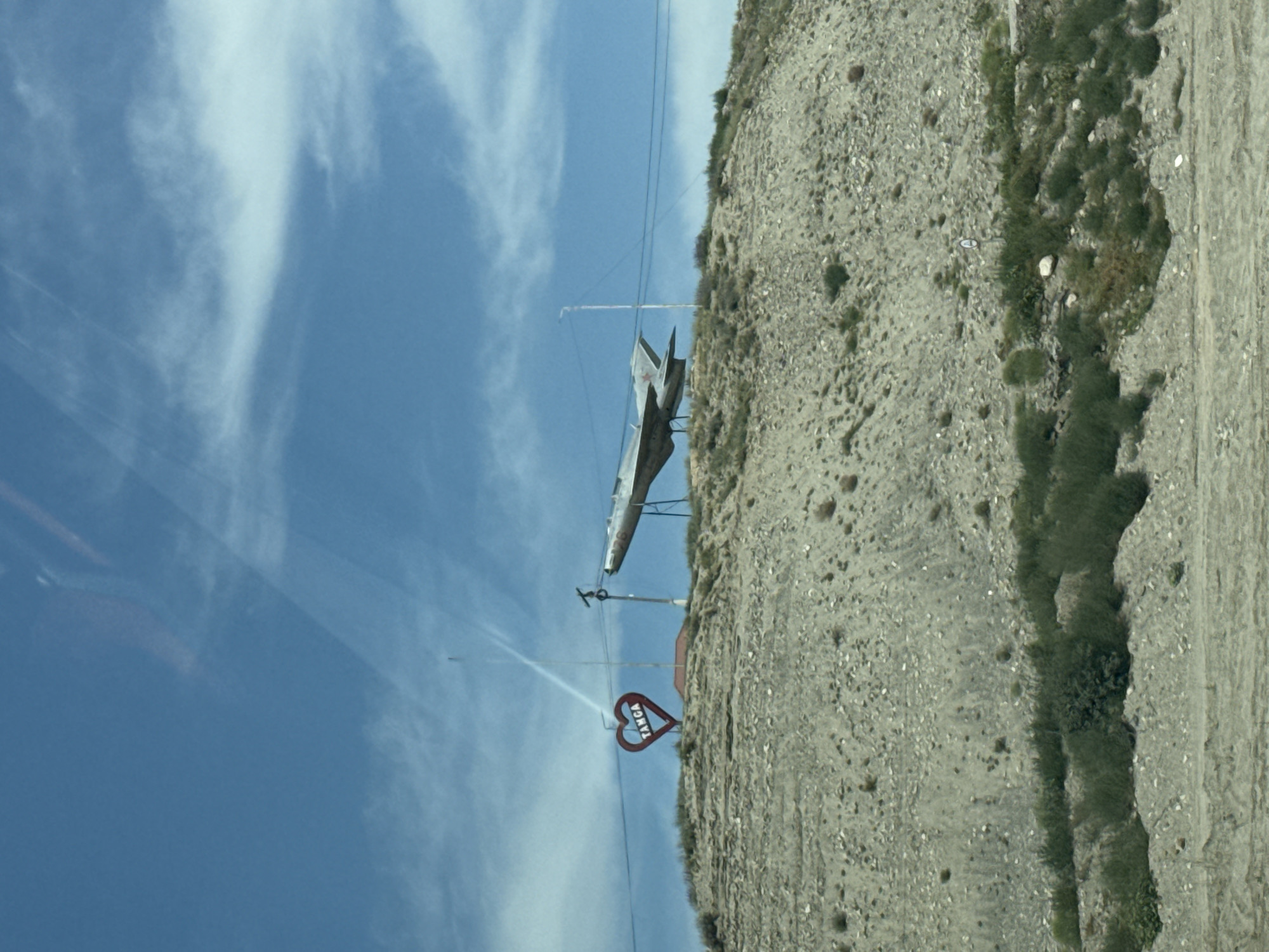 The image shows a hillside in Tamga, Kyrgyzstan, featuring a large metallic sculpture of a fighter jet mounted on a stand. To the left of the jet, a red heart-shaped sign with the word "TANGA" inscribed in white lettering is visible. The hillside is sparsely covered with dry grass and rocks. Power lines and a tall pole with a light are also present in the scene. The sky is blue with scattered white clouds. The overall impression is that of a roadside attraction or monument in a rural or semi-arid environment.