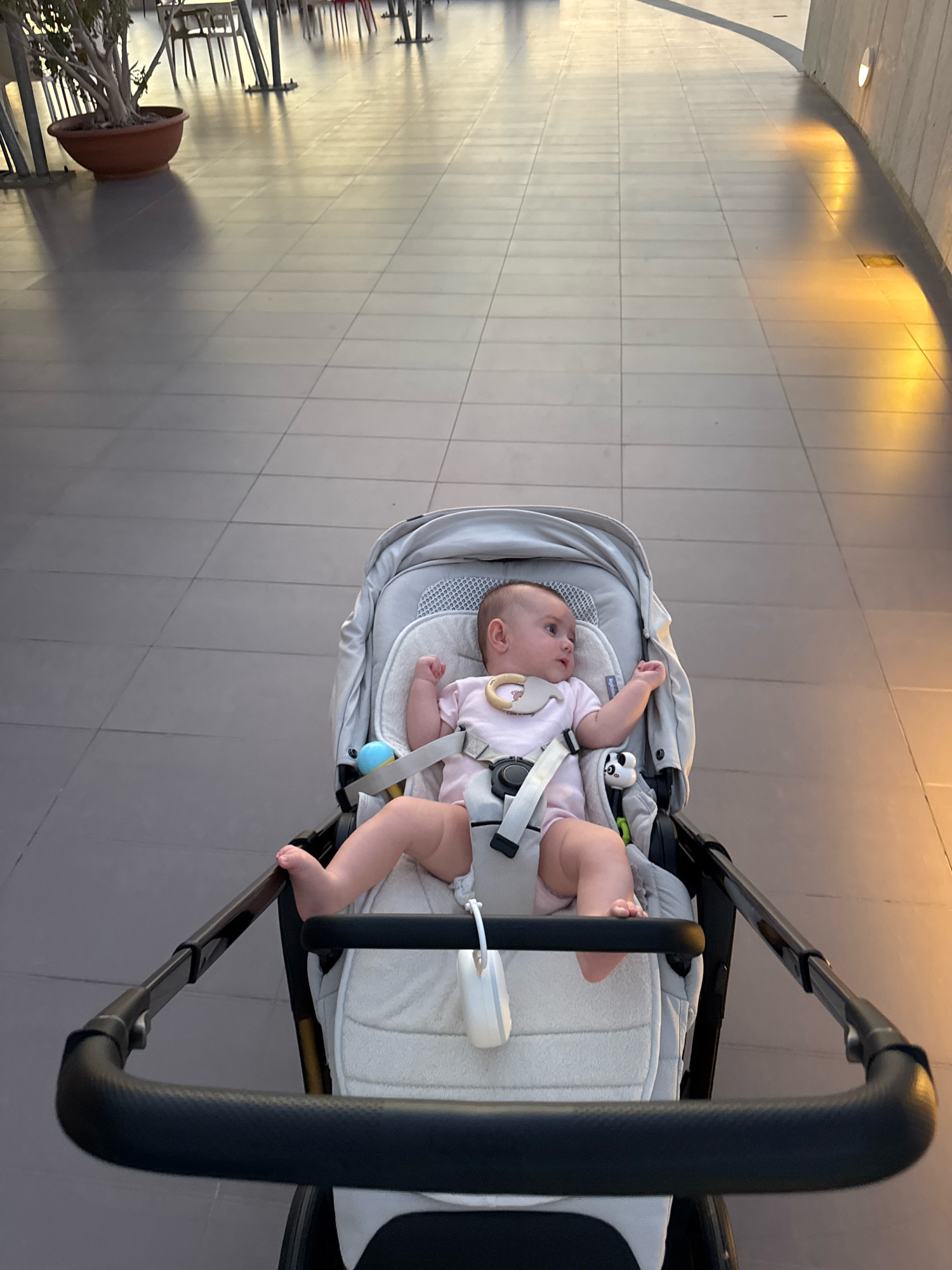 An overhead shot shows a baby in a stroller on a tiled walkway. The baby is wearing a pink outfit and is strapped into the stroller. The stroller has a gray frame and light-colored padding. In the background, there is a potted plant and some tables and chairs. The walkway is illuminated by soft, warm lighting, casting long shadows on the ground. The scene appears to be an outdoor public space, possibly a promenade or a plaza, with modern architecture visible on the right side of the image. The overall impression is one of a peaceful, daytime outing. The location is Isola delle Femmine, Italy.