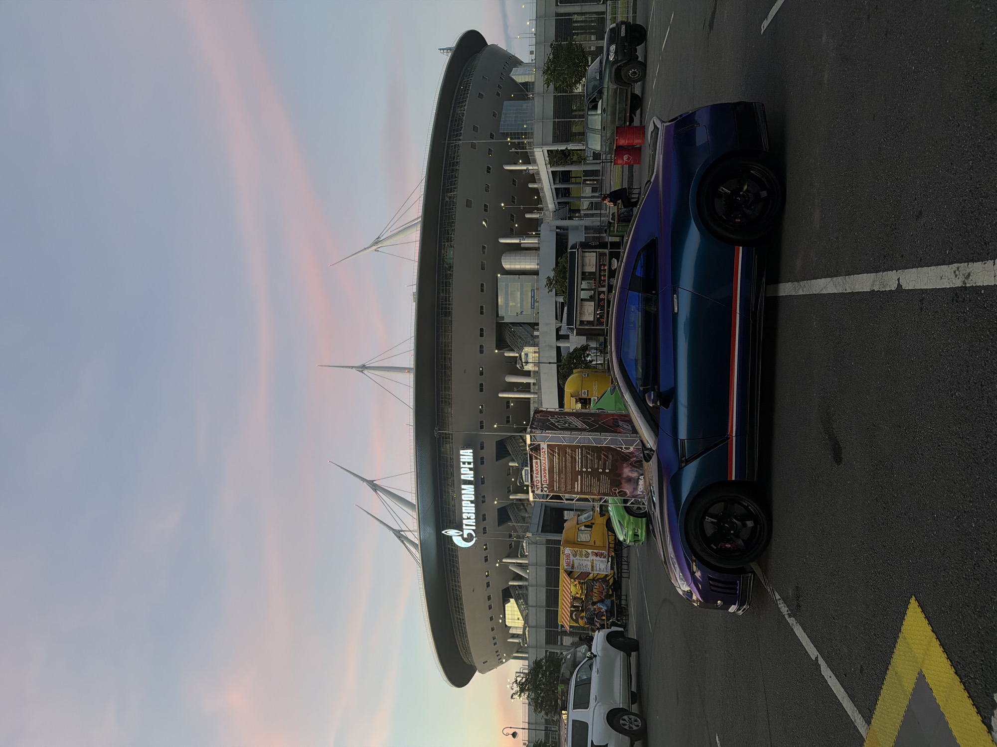 The image captures a twilight scene in an outdoor parking lot in front of the Gazprom Arena. The sky displays a soft gradient of blue and pink, indicative of sunset. A striking, iridescent blue and purple sports car, likely a Nissan GT-R, is prominently parked in the foreground, facing right, with its wheels turned slightly. The car features a sporty body kit and red and white racing stripes along its side.
Behind the sports car, several other vehicles are visible: a white SUV to the left, a green hatchback, and a darker-colored off-road vehicle to the far right. A food truck with a yellow awning is parked behind the white SUV. In the background, the Gazprom Arena stadium stands as a dominant structure, illuminated by bright lights. The stadium's distinctive architecture, with its sharp white spires, is clearly visible against the sky. A large banner with Russian text and imagery is also present, situated between the food truck and the off-road vehicle. The overall environment suggests a public gathering or event space.