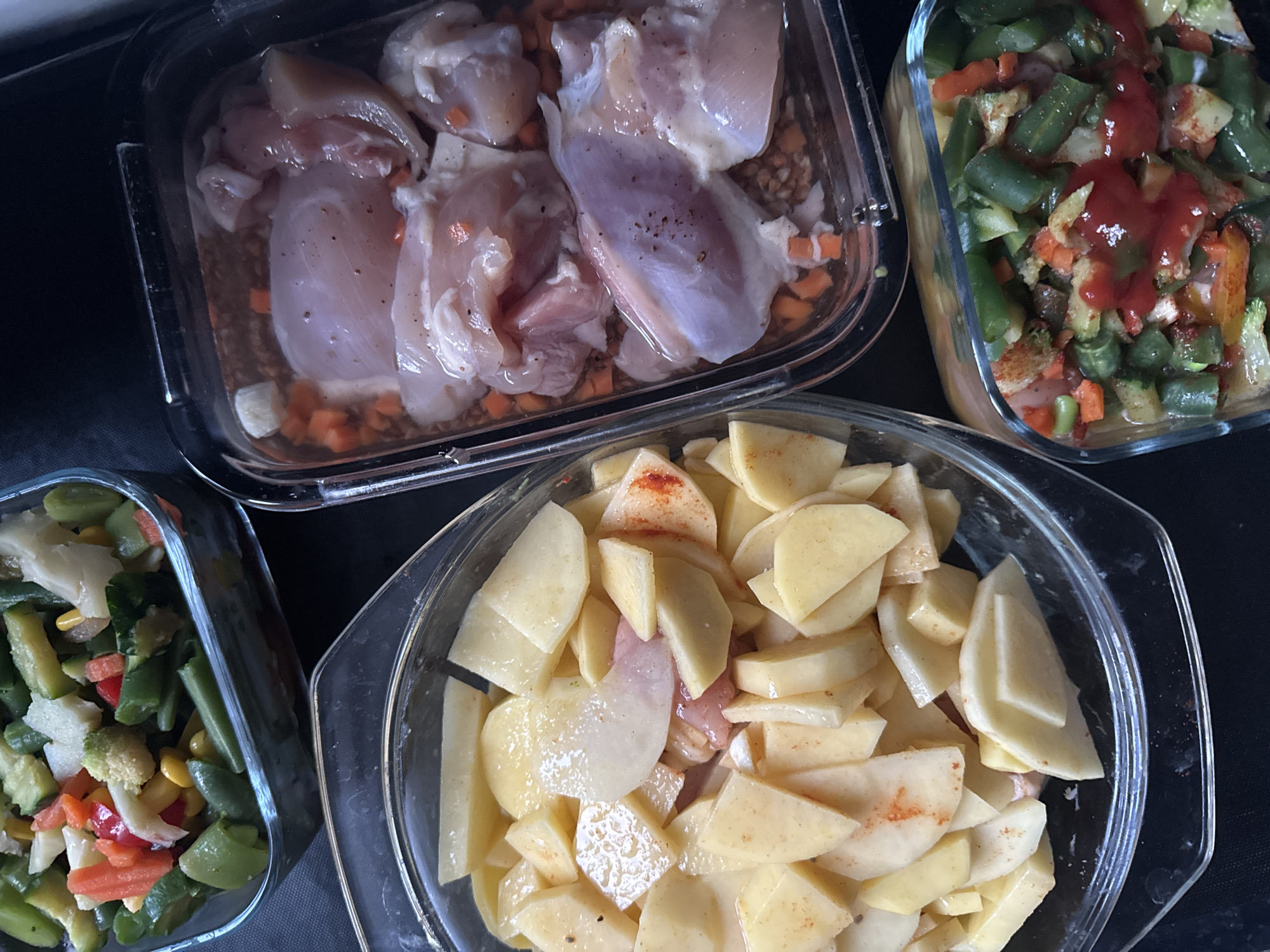 The image shows four glass dishes containing food.
The dish in the upper left contains raw chicken pieces in a marinade with diced carrots. The dish in the upper right contains cooked green beans, carrots, and what appears to be tomato sauce. The dish in the lower left shows mixed vegetables, including green beans, corn, and carrots. The dish in the center contains sliced potatoes sprinkled with a red spice, possibly paprika, with a few pieces of chicken underneath. The background is a dark, possibly black, surface. The scene is indoors.