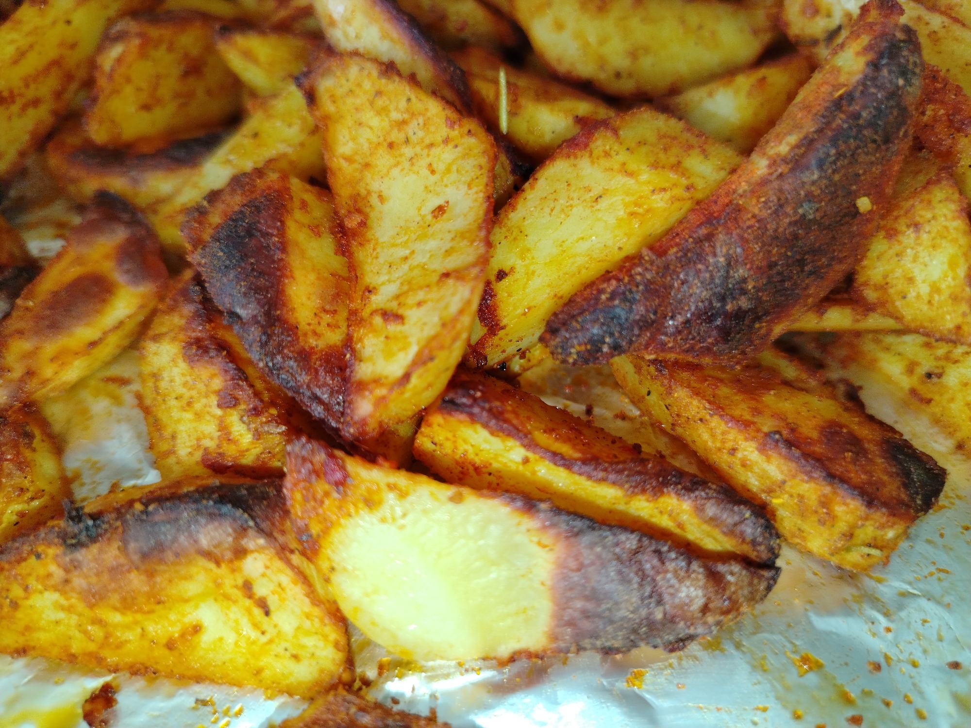 The image shows a close-up of roasted potato wedges. They are heavily seasoned with a reddish-orange spice and appear to be slightly overcooked, with some edges darkened or slightly burnt. The potatoes are arranged on a surface covered with aluminum foil. There are no people or indications of a specific location like Noviy, Russia visible in this image.
