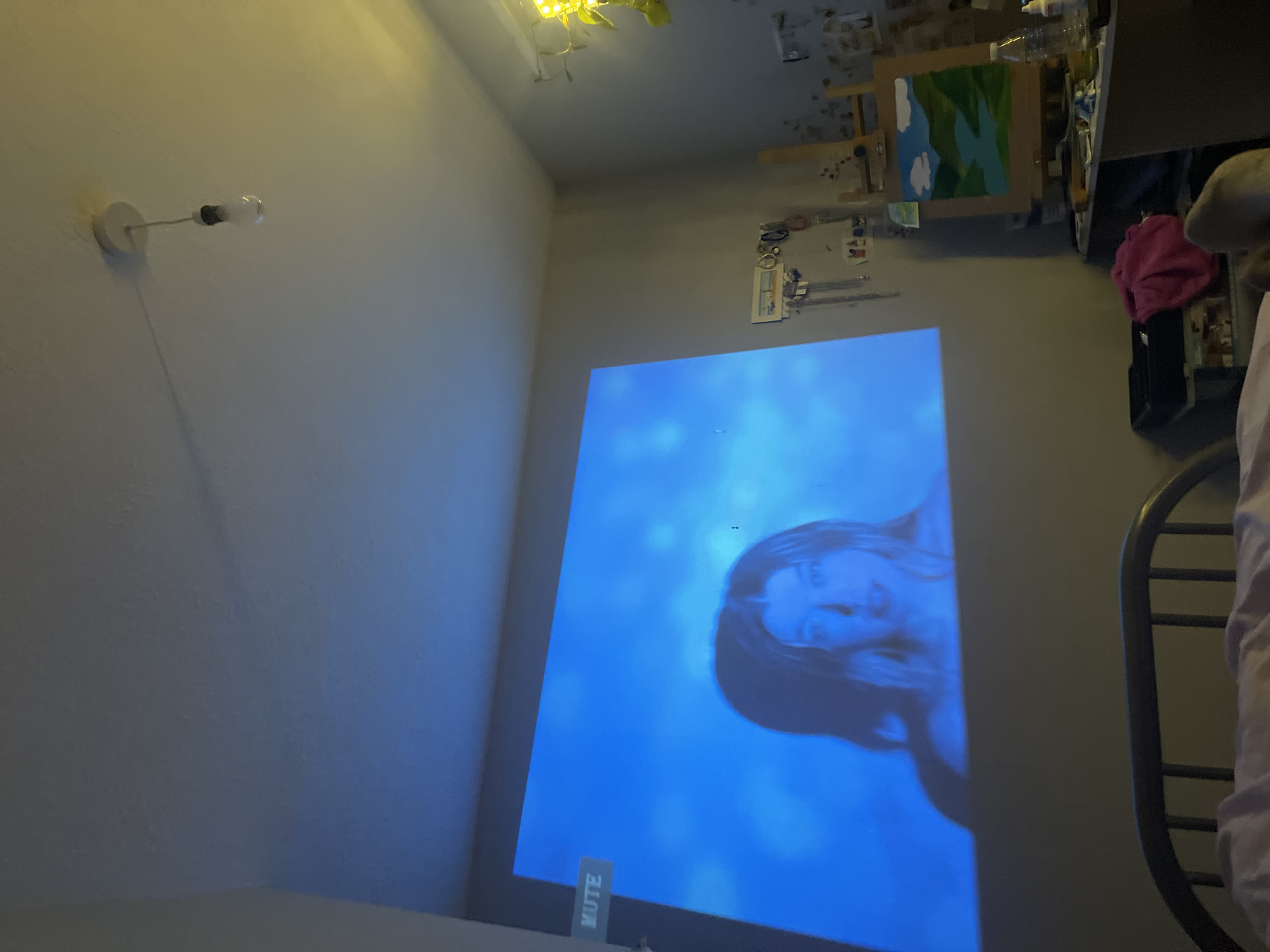 A photograph taken indoors features a wall projection displaying a human face in blue tones. Adjacent to the projection, a metal chair is partially visible. In the upper right corner, a painting on an easel stands near various containers and a pink garment. A light fixture is located on the upper left wall. The photograph was taken at an unknown location in Maidstone, United Kingdom.