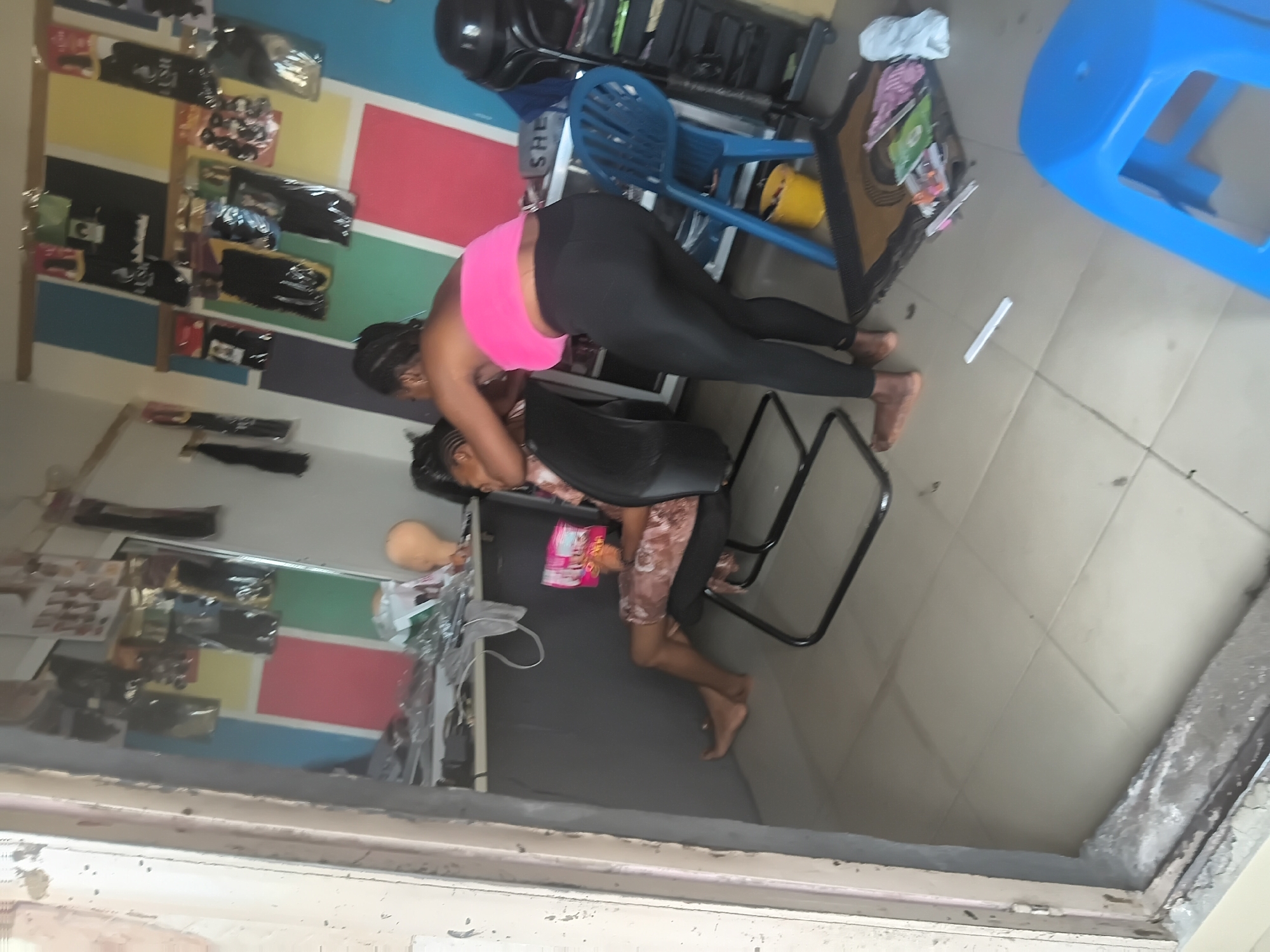 This image presents the interior of a hair salon in Jalingo, Nigeria, observed through an opening in the immediate foreground. Two adult females are present. One woman, wearing a bright pink tube top and black leggings, is standing and actively styling the hair of a second woman. The standing woman is barefoot. The second woman is seated in a black office chair, wearing a light-colored, patterned short dress, holding a pink package, and looking downwards. This woman is also barefoot.
The rear wall of the salon is adorned with large vertical stripes in alternating blue, yellow, pink, and green sections, against which numerous packaged hair extensions and products are displayed. Below the wall display on the left, a dark desk or counter holds various items, including a light-colored mannequin head. To the right, a black hooded hair dryer on a stand is visible next to a dark, multi-tiered shelving unit containing hair products. A blue plastic stool, a yellow bucket, a black floor mat, and additional items such as packaged products and a white cylindrical object are situated on the light-colored tiled floor. The scene depicts an active small-scale hair styling business.