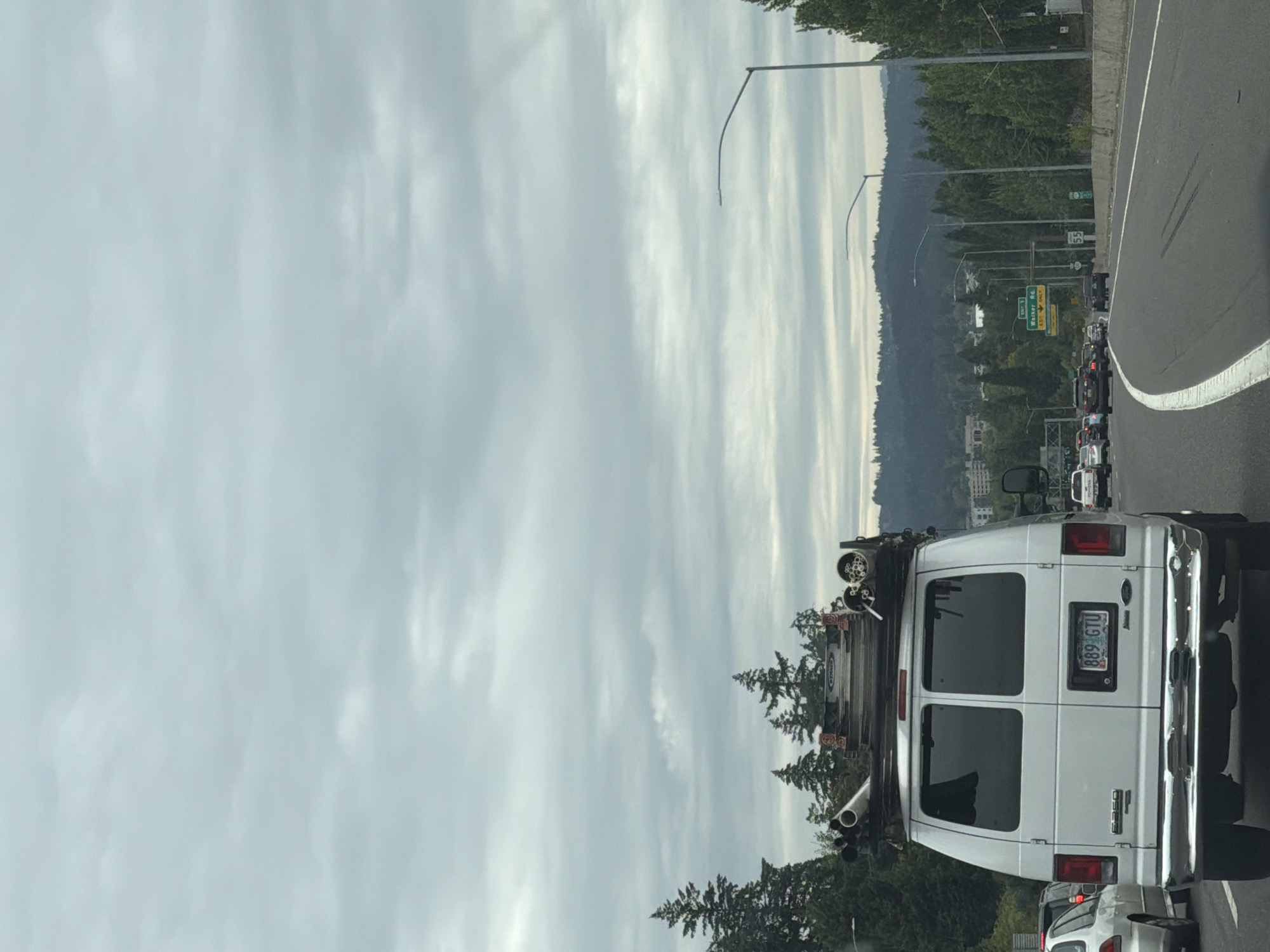 This is a photograph taken from inside a vehicle, looking out at a cloudy sky and a highway. The image is oriented vertically. In the foreground, the rear of a white van is visible. The van has a ladder rack on top with several pipes and other equipment. The license plate on the van reads "889 GTU". Behind the van, other vehicles are seen in traffic. The highway curves to the right, with a concrete barrier and trees lining the side. In the distance, a densely wooded hillside rises, with some buildings visible among the trees. Streetlights are present along the highway. Several road signs are visible, including one that says "EXIT 1" and another with the number "55". The sky is overcast with streaks of light suggesting it is either early morning or late afternoon. The scene appears to be a typical highway commute in a suburban or semi-rural area. There are no people prominently featured in the image. The overall mood is mundane, depicting everyday travel.