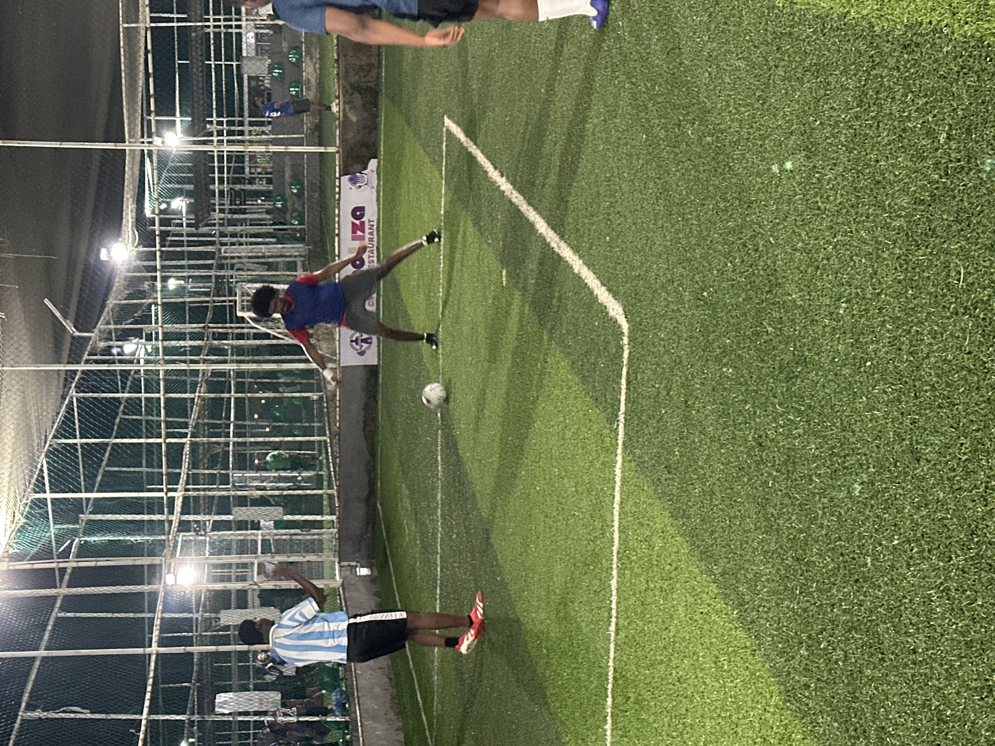 A group of young men are playing soccer on an artificial turf field at night in Abuja, Nigeria. The field is enclosed by a white net fence and illuminated by stadium lights, creating a nighttime playing environment. The men are dressed in athletic attire, with some wearing jerseys and shorts. One man in a blue and red jersey is in the process of kicking a soccer ball. Another man in a blue and white striped jersey stands to the left, observing the game. In the background, spectators can be seen watching from behind the fence. The scene suggests a recreational or competitive soccer match taking place in an urban setting.