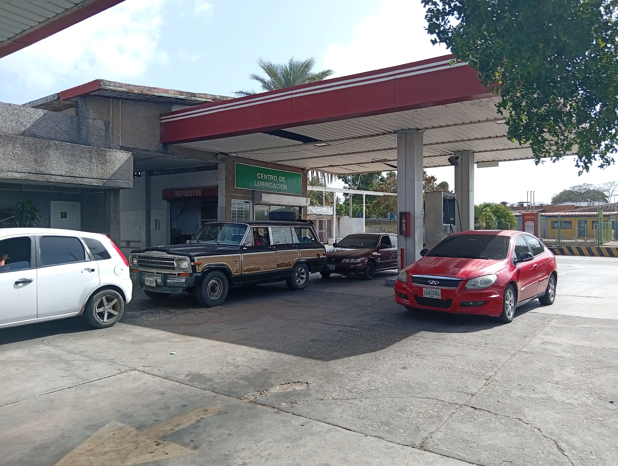The image depicts a gas station in La Tagua, Venezuela. Visible are several vehicles: a white hatchback, a black and tan Jeep Wagoneer with a spare tire on its roof, a dark red sedan, and a red hatchback. The gas station has a red and white canopy supported by white pillars. A sign reads "CENTRO DE LUBRICACION." To the left of the canopy is a building with a sign that reads "REPUESTOS." The ground is concrete with visible cracks and stains. In the background, there are buildings and trees. A palm tree is visible above the canopy. The sky is clear and sunny.