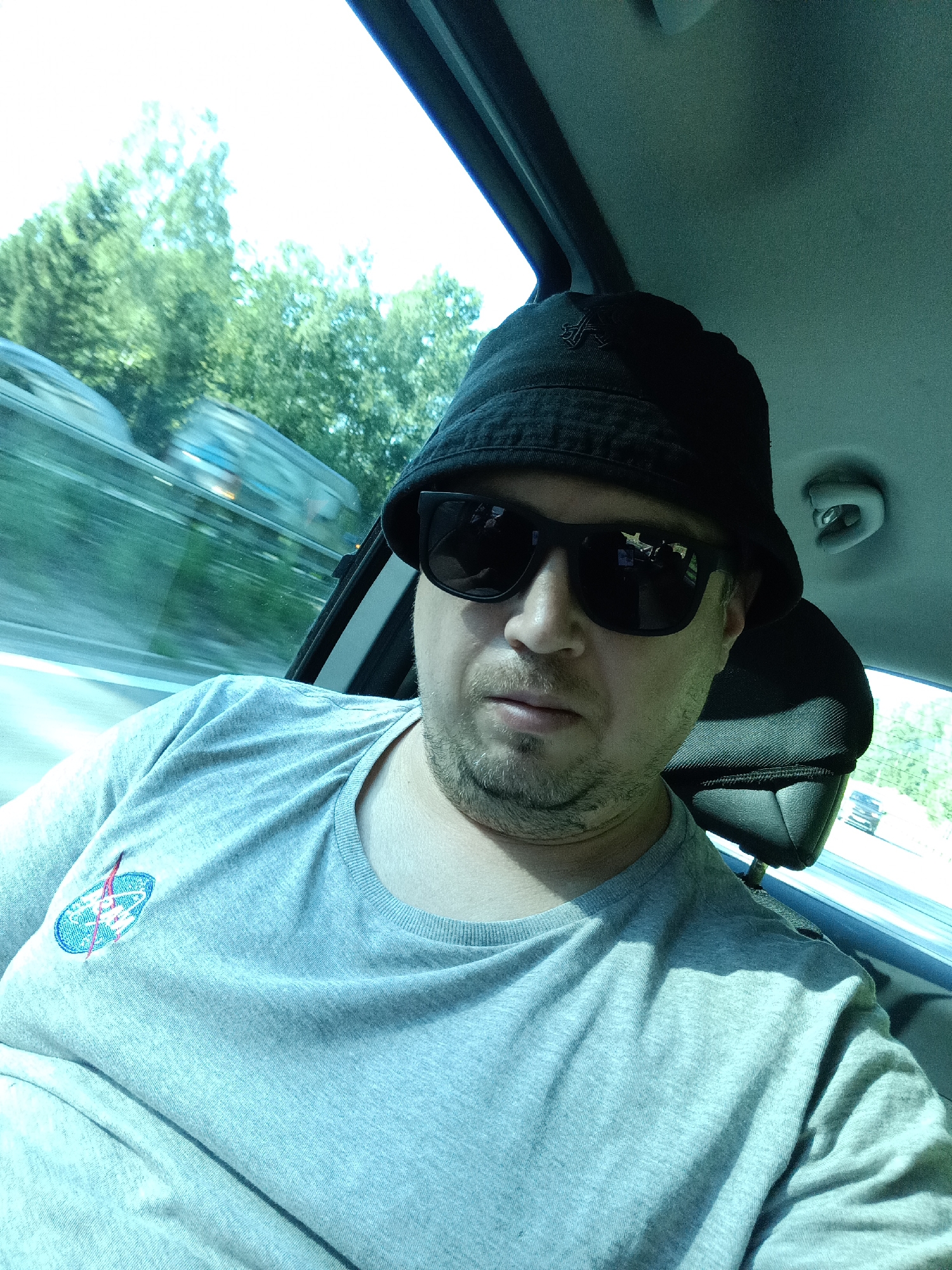A man wearing a black bucket hat and dark sunglasses is sitting in a car, taking a selfie. He is wearing a light gray t-shirt with a NASA logo embroidered on the left chest. The car appears to be in motion, as indicated by the blurred trees and road visible through the windows. The scene is outdoors, with bright sunlight and green foliage visible. The image is a selfie taken by the man. The location is stated as Psáře, Czechia, however, there are no identifying features in the image to confirm this.