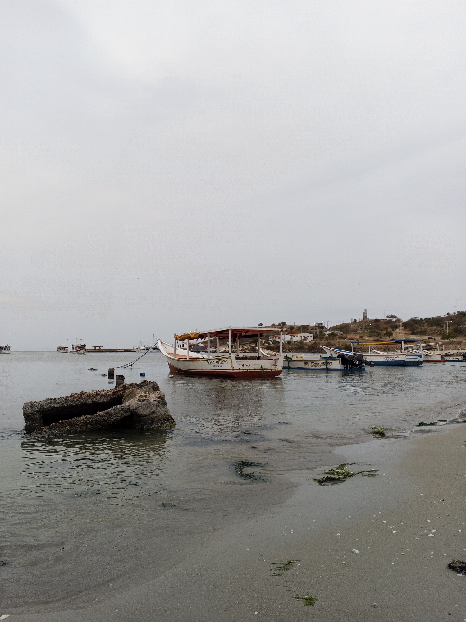 The image depicts a coastal scene in Juan Griego, Venezuela, under an overcast sky. In the foreground, a partially submerged, broken concrete structure rests in the shallow water near a sandy beach strewn with seaweed. Several boats are moored further out in the bay, with the closest one being a white and red boat bearing the name "Marc Anthony." Additional boats are visible along the shoreline in the background. On the right side of the frame, a hillside dotted with sparse vegetation and a few buildings rises above the bay. In the distance, a tall, light-colored structure stands on the crest of the hill, possibly a monument or tower.