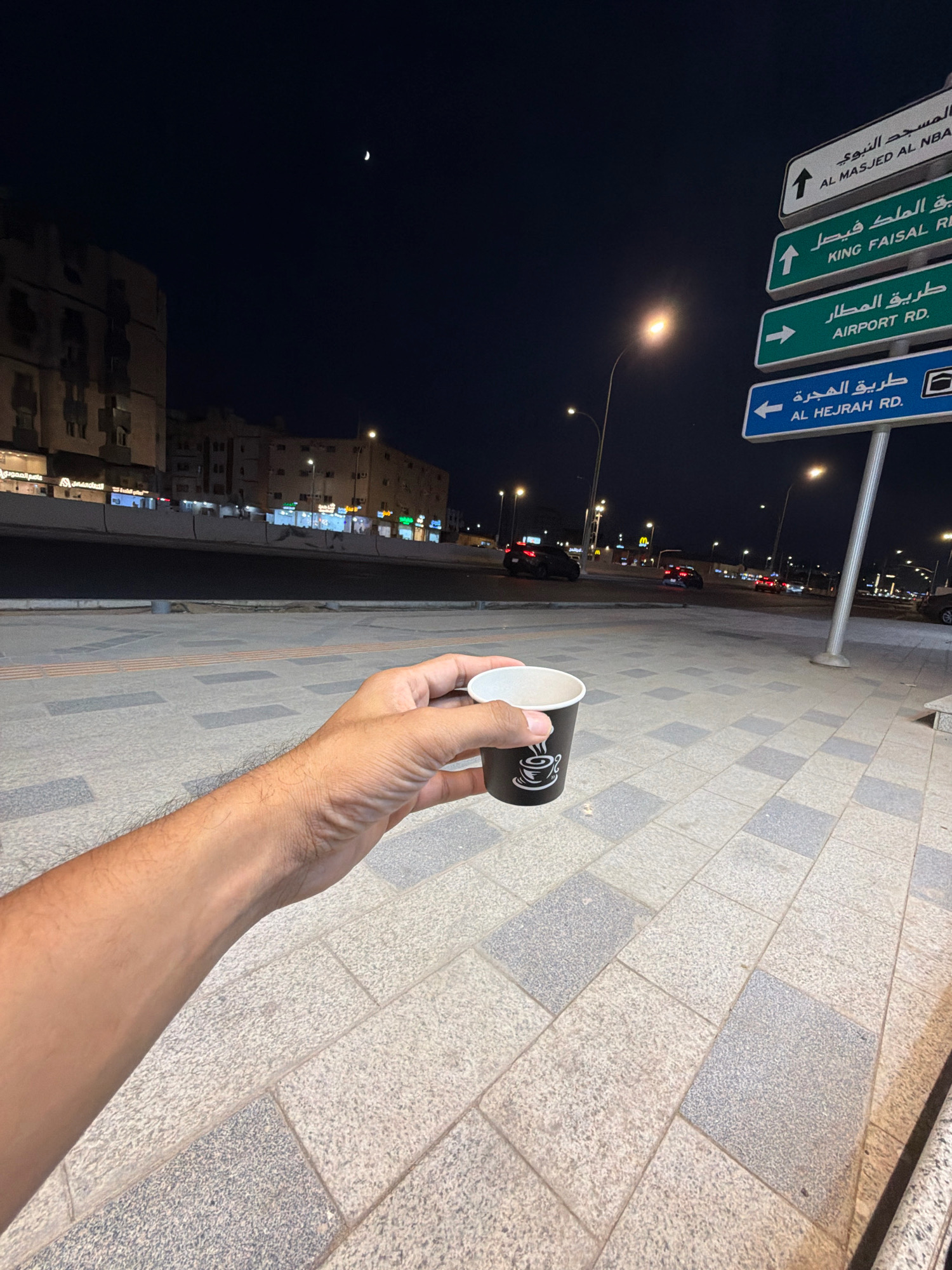 A first-person perspective shot captures a hand holding a small, black and white paper coffee cup with a coffee cup graphic on it. The hand is on the right side of the frame, with the arm extending leftwards and downwards. The cup is positioned near the lower right corner of the image. In the background, a wide, paved walkway with a gray and tan patterned tile design stretches into the distance. Beyond the walkway, a street is visible at night, with several cars driving and streetlights illuminating the scene. Buildings line the street on the left side of the frame, with illuminated windows and signs. A tall signpost with multiple green and white directional signs written in Arabic and English stands on the right side of the image. The signs indicate directions to "AL MASJED AL NBA", "KING FAISAL RI", "AIRPORT RD", and "AL HEJRAH RD". The night sky above is dark, with a faint crescent moon visible in the upper left quadrant. The overall scene depicts an urban environment in Medina, Saudi Arabia, at night.