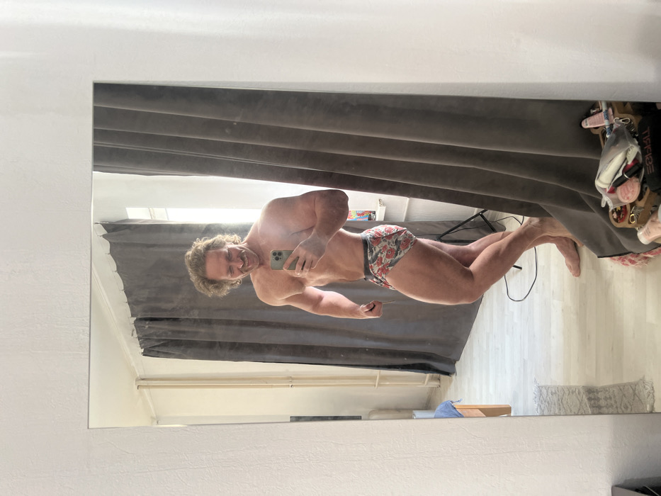 The image displays a muscular, fair-skinned man with curly blond hair and a prominent mustache, smiling broadly as he photographs his reflection in a large, rectangular mirror with a white frame. The scene is located within a room in Saint Petersburg, Russia. The man is shirtless, wearing floral-patterned boxer briefs featuring red flowers and green foliage on a light background, and is barefoot. He holds a light-colored smartphone in his right hand, positioned to capture the mirror selfie, while his left arm is slightly flexed, and his body is angled to showcase his physique.
The reflected environment behind the man reveals a room with light-colored walls and a light wooden floor. To the man's right, floor-to-ceiling dark grey curtains are partially drawn, revealing a window with a white frame and bright natural light filtering in. A vertical white pipe or radiator runs alongside the window. To the man's left, additional dark grey curtains are visible, along with a black photographic light stand positioned behind his lower left leg. On the far left of the reflection, part of a wooden bed frame with a blue pillow can be seen. In the immediate foreground, outside the primary reflected image, various objects including makeup items (lipstick, compact) and other small containers are visible on a surface in the bottom right corner of the mirror's actual frame.