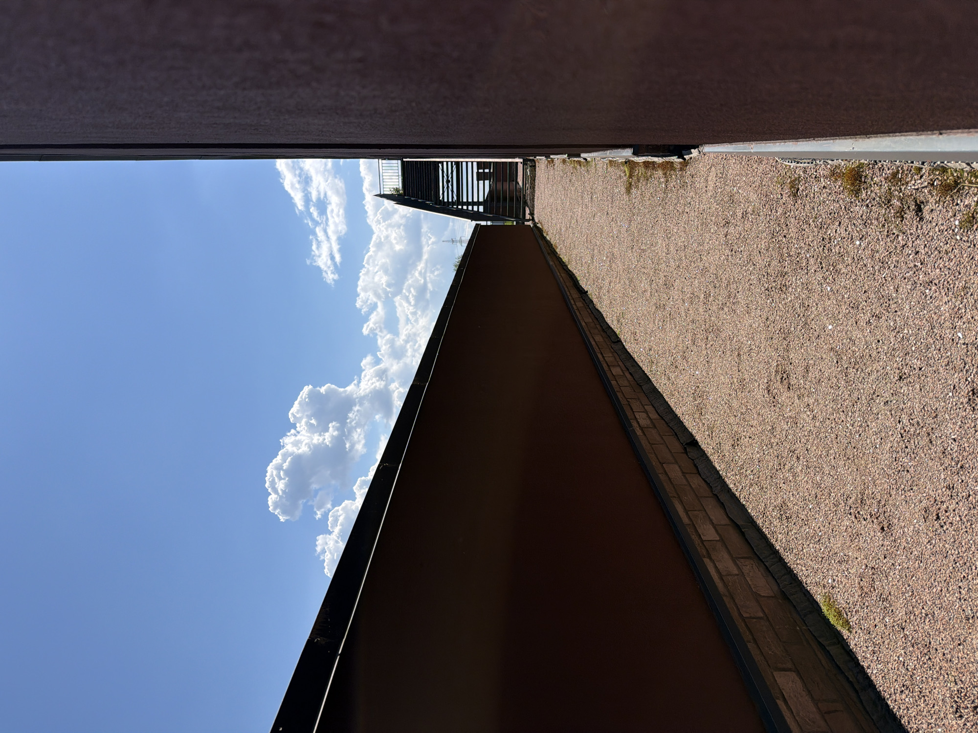 The image captures a narrow, linear outdoor space, viewed in a vertical orientation. On the left side, a vibrant blue sky is visible, populated by scattered, fluffy white cumulus clouds. The right side of the frame is dominated by a light brown, gravel-covered path or ground surface, which extends into the distance.
Two long, dark brown or rust-colored structures frame the scene horizontally. One runs along the top edge of the frame, acting as an overhead canopy or wall. The other forms a low wall or barrier along the bottom edge, bordering the gravel path. Both structures appear solid and possibly made of weathered metal or a similar robust material. In the far distance, towards the upper right, a dark, multi-level metallic structure resembling an outdoor staircase or fire escape is visible, through which the sky and clouds can be seen.
The scene is static, devoid of people or discernible activity, presenting an architectural landscape.
Notable details include the bright, sunny daytime lighting, indicated by the clear blue sky, bright clouds, and distinct shadows. Small patches of green moss are visible along the edge where the gravel path meets the lower dark wall. The structures suggest an industrial or minimalist architectural style, creating a stark contrast with the natural elements of the sky and gravel. There is no visible text in the image.