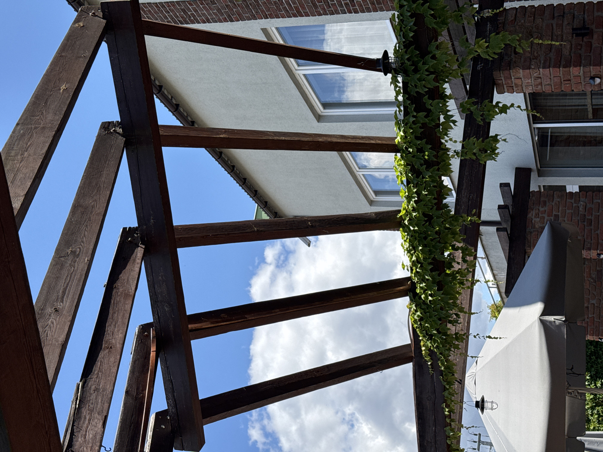 An outdoor scene beneath a wooden pergola covered in green ivy. The pergola's dark wooden beams are visible against a bright blue sky with scattered white clouds. Beyond the pergola, a light-colored building with several white-framed windows and a red brick chimney is visible. In the foreground, a portion of a large, light grey umbrella is present. This image was taken in Duszniki, Poland.