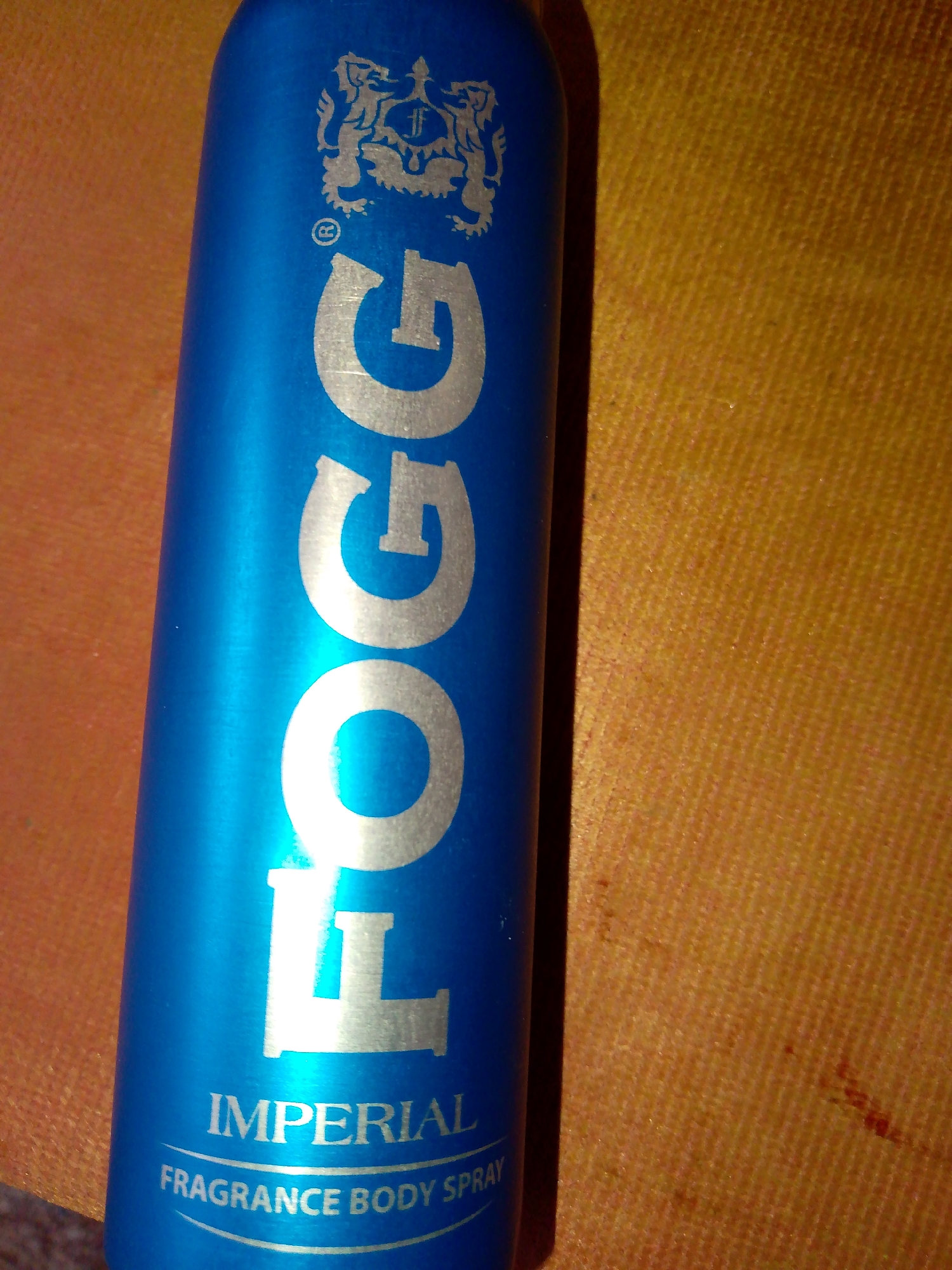 An upright blue aerosol can is positioned diagonally on a textured orange surface. The can has silver lettering and a logo. The word "FOGG" is written vertically in large, stylized silver letters on the can. Above the word "FOGG", a silver logo is visible, depicting a crest with two lions and an 'F' in the center. Below the word "FOGG", the word "IMPERIAL" is written in smaller silver capital letters, followed by the words "FRAGRANCE BODY SPRAY" in even smaller silver capital letters, arranged in a curved line. The orange surface appears to be fabric, possibly a tablecloth or a textured wall. The lighting casts a shadow from the can onto the orange surface, and some parts of the can are highlighted by a light source. There are no people or other objects in the frame. The setting is indoors, with no discernible environmental features indicating a specific city or location.