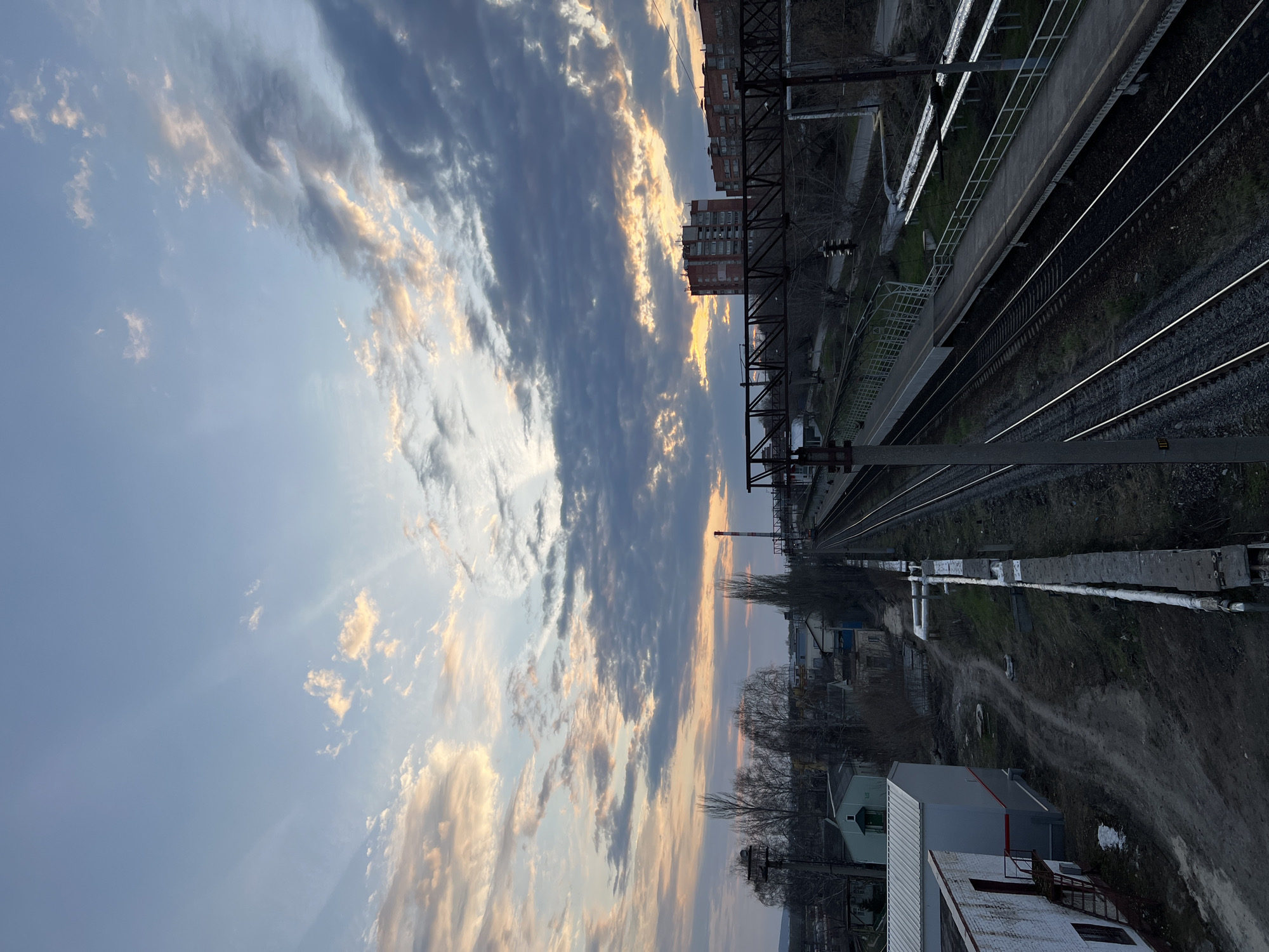 The image depicts a scene from Kazan, Russia, featuring a railway track running through an urban area. The railway is a central element, with parallel tracks and supporting infrastructure like poles and wires extending into the distance. A train platform runs parallel to the tracks, with railing to mark the edge. Adjacent to the railway, there are industrial or residential buildings, showing the proximity of the rail line to urban structures. The sky is filled with clouds. The view is captured from an elevated perspective, potentially from a higher building or structure overlooking the railway. The lighting suggests either sunrise or sunset. The environment surrounding the railway appears to be a mix of industrial and residential areas.