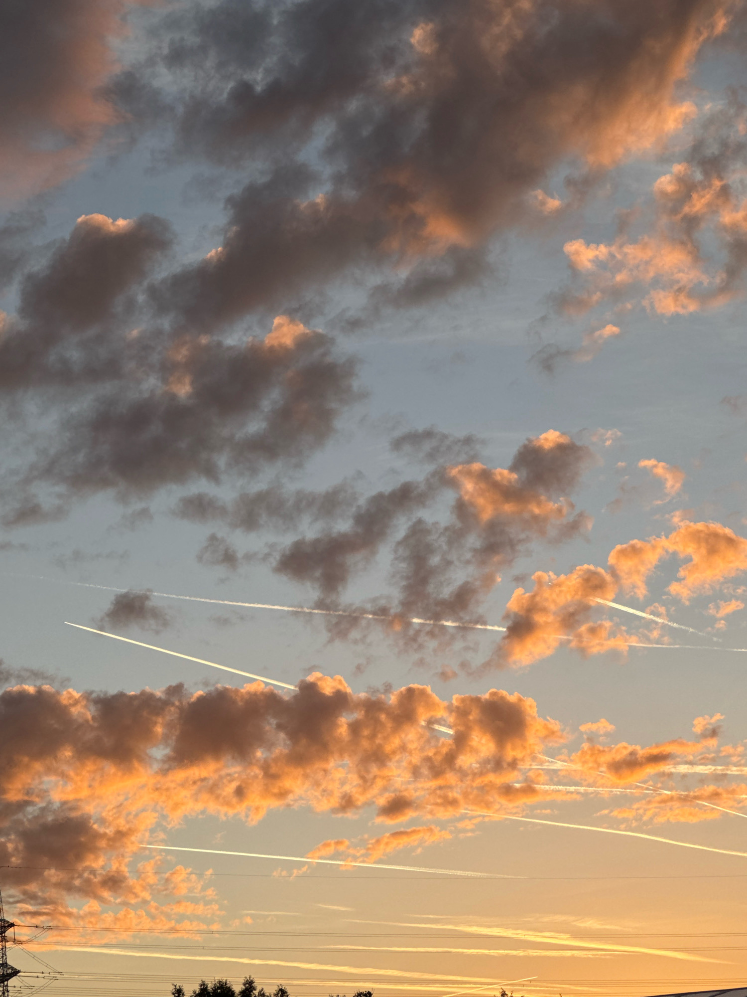A dramatic sunset illuminates a sky filled with a mix of dark, wispy clouds and brighter, orange-tinted clouds over Wettstetten, Germany. Multiple white contrails from aircraft crisscross the sky, creating linear patterns against the colorful backdrop. The lower portion of the image shows a silhouetted treeline and overhead power lines, indicating a partly urban or suburban setting. The lighting suggests it is late afternoon or early evening.