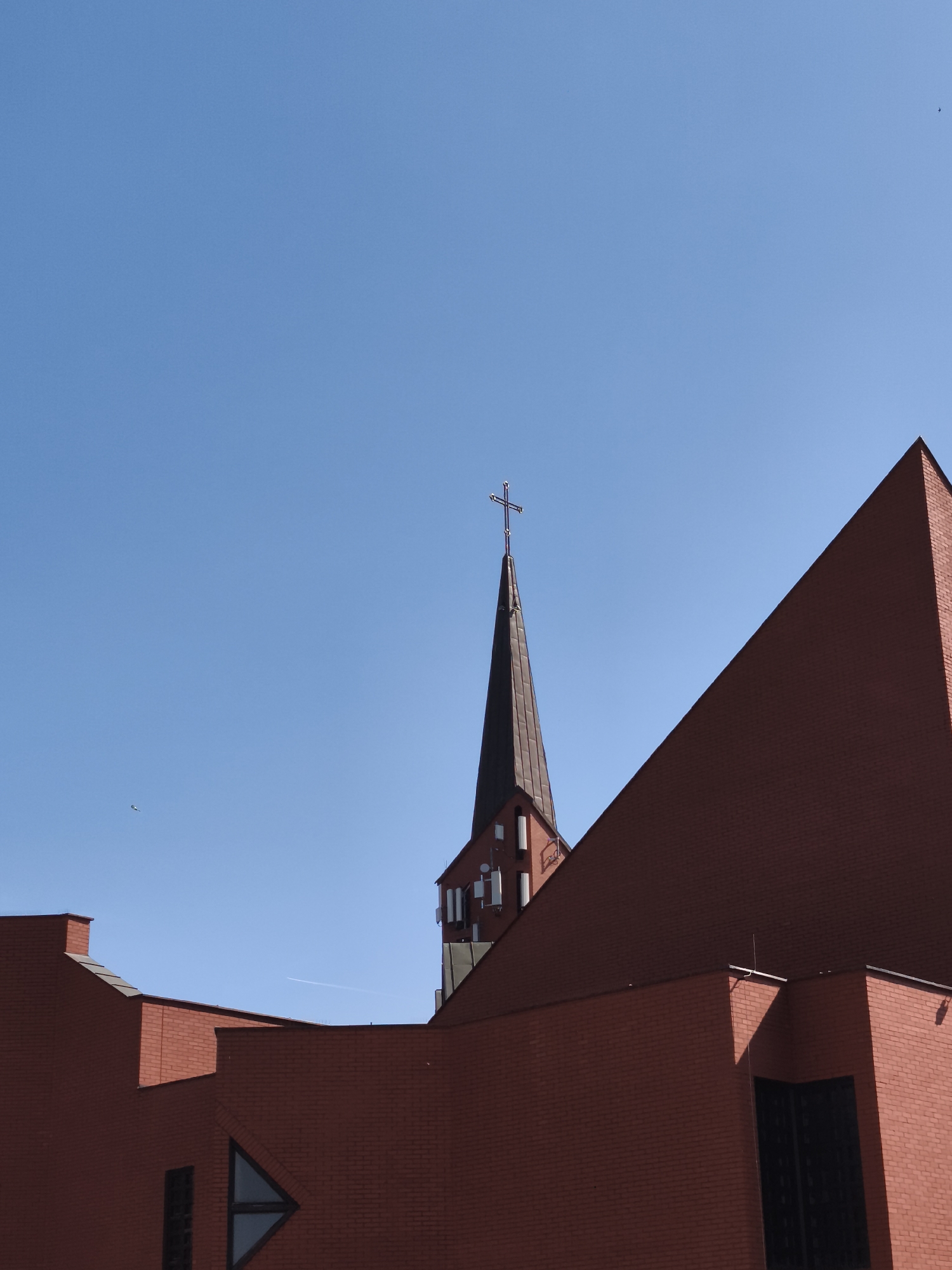 The image shows the St. Mary's Church in Gdańsk, Poland, under a clear blue sky. The church is constructed of red brick and features a tall, pointed spire topped with a cross. The foreground is dominated by the angled brickwork of the church's roof and walls, with visible windows and architectural details. There are a few distant birds and a faint contrail in the sky, suggesting an open, expansive environment. The lighting indicates it is daytime, with strong shadows cast by the sun on the building.