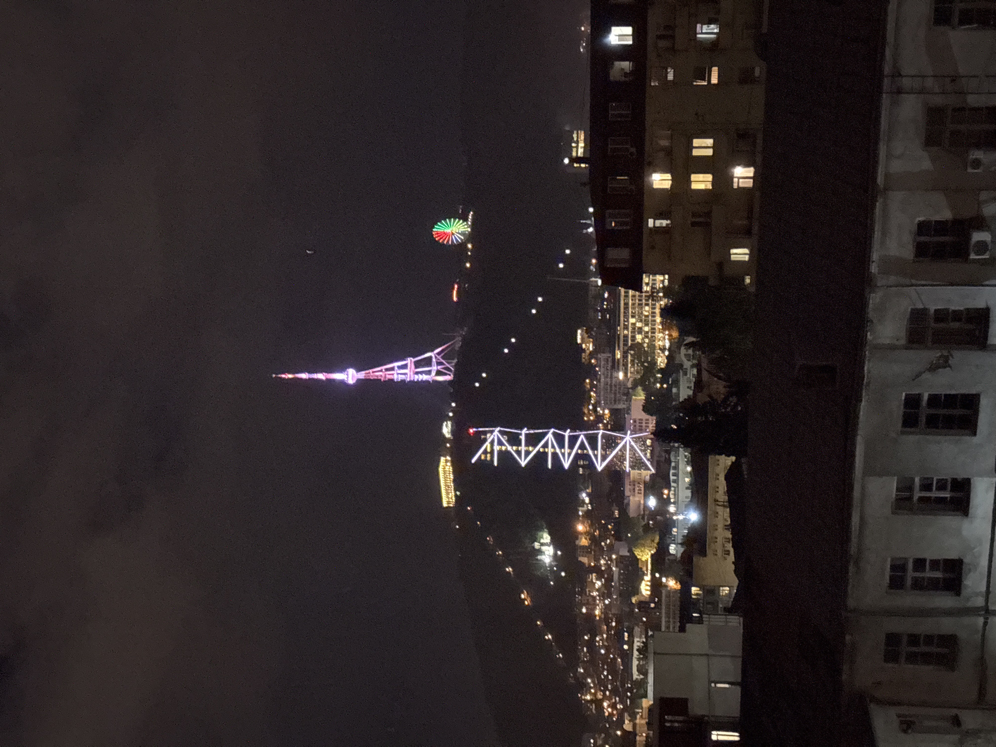 The image shows a night scene in Tbilisi, Georgia. Visible in the distance are the illuminated Tbilisi TV Broadcasting Tower, the Bridge of Peace, and a Ferris wheel. Buildings line the right side of the image, with windows illuminated. The sky is dark, with some cloud cover. The city lights stretch into the distance, indicating the urban sprawl.