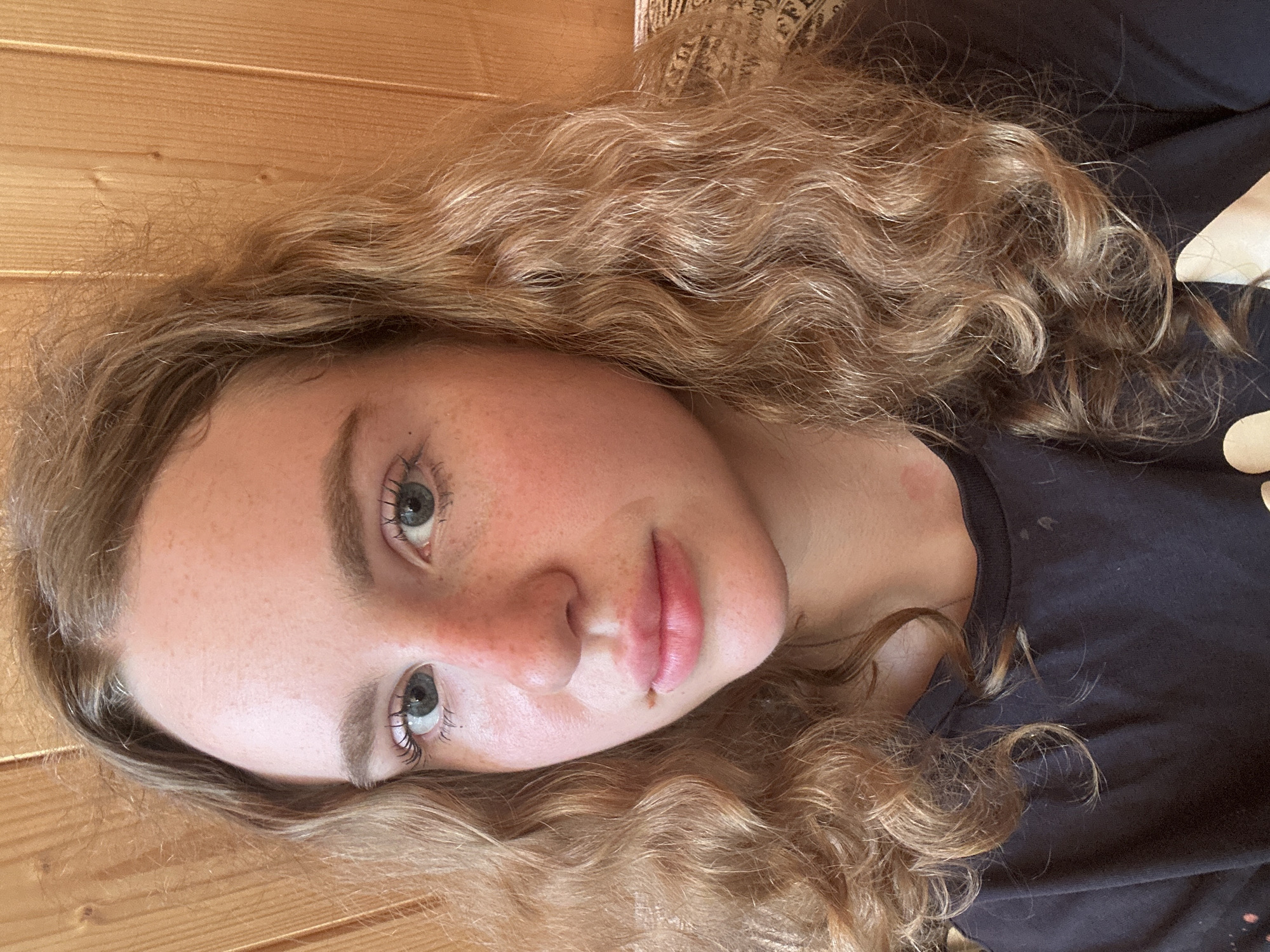 A medium close-up selfie of a young woman with blonde, wavy hair, freckled skin, and blue eyes. She is wearing a dark t-shirt and her head is tilted slightly to her left. The background is composed of vertical wooden planks. The image captures a natural pose and lighting, suggesting an indoor or outdoor setting with diffused light. No specific location in Vasilyevo, Russia is discernible from the image content.