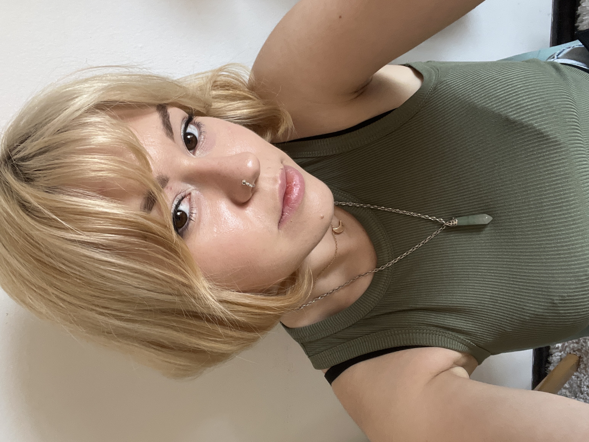 A woman is lying on a white surface. She has blonde hair, brown eyes, and a nose ring. She is wearing a green tank top with a black sports bra underneath. She has two necklaces: one with a crescent moon pendant and the other with a green crystal pendant. The location is unknown, as there is nothing to indicate the city of Budva, Montenegro.