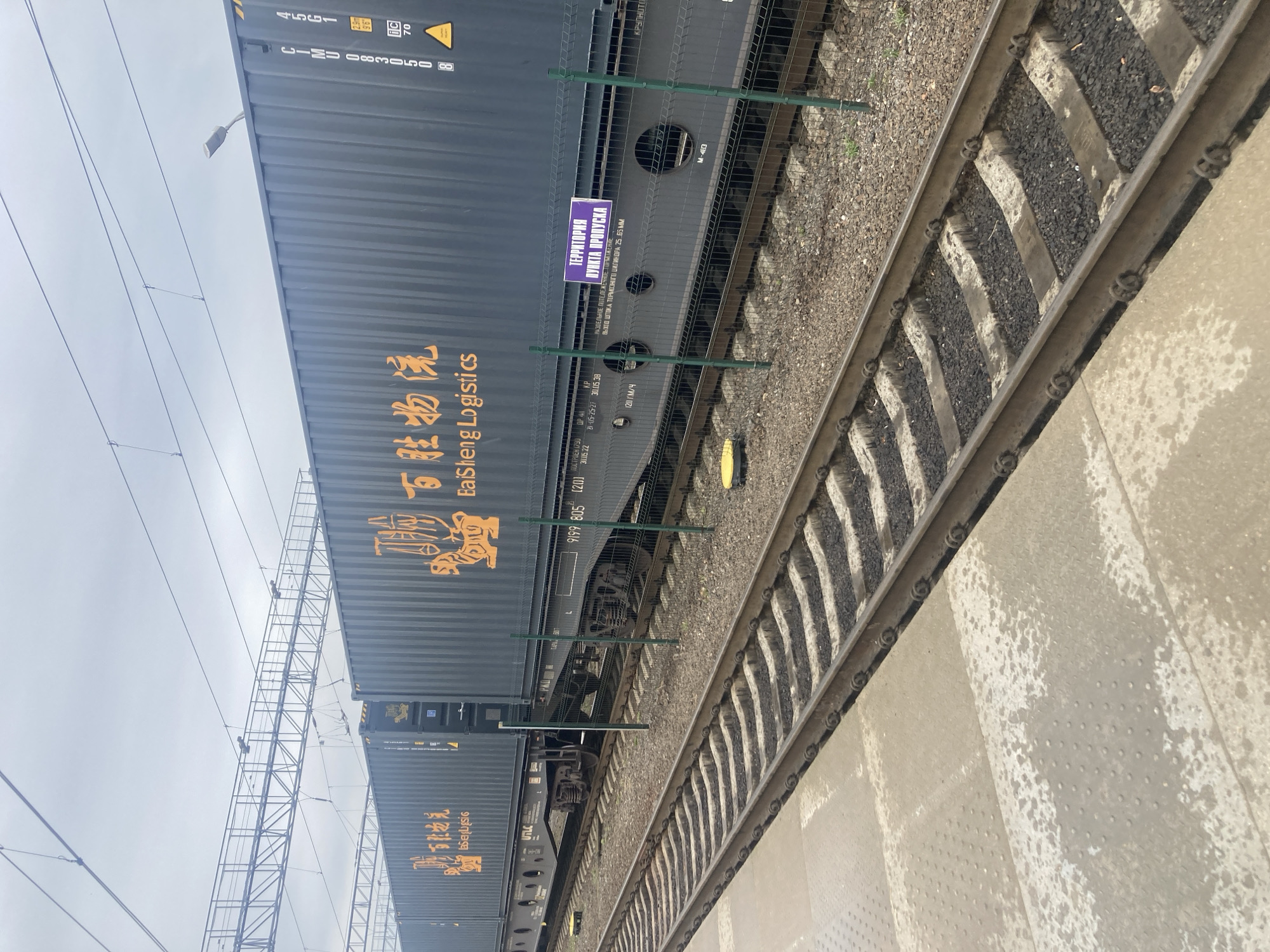 The image shows a train stopped at a station in Brest, Belarus. The train consists of several freight containers, each marked with orange Chinese characters and the text "taSheng Logistics". On one of the containers, a purple sign reads "ТЕРРИТОРИЯ ПУНКТА ПРОПУСКА" in Cyrillic, which translates to "PASSAGE POINT TERRITORY". The train is on tracks, with a platform visible in the foreground. Overhead, electrical wires for the train are suspended from a metal structure. The sky is overcast.
