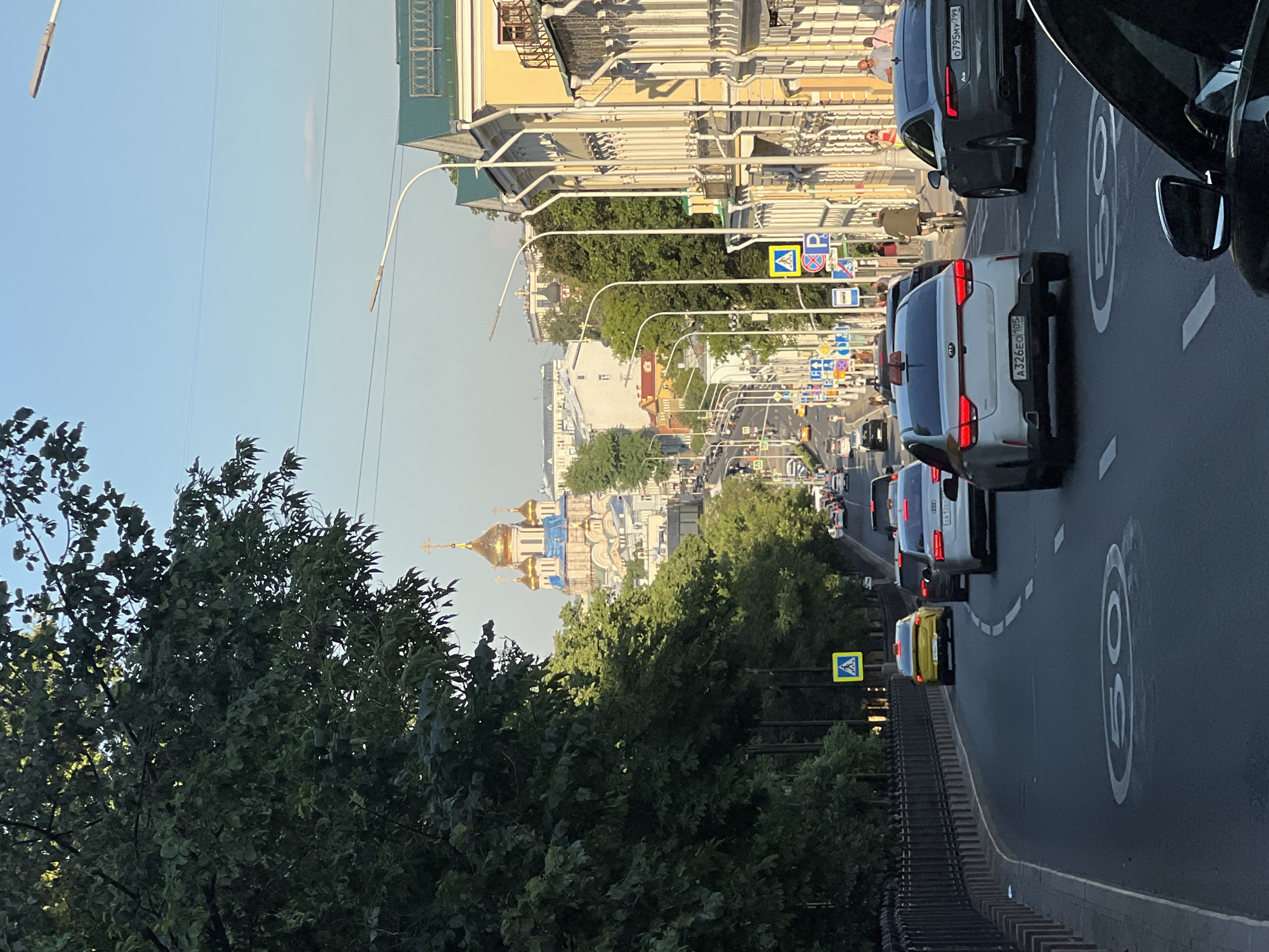 The image shows a street scene in Moscow, Russia, looking down a hill towards a golden-domed church in the distance. The street is lined with trees and buildings on the right. There are several cars on the road, including a yellow Kia, a white Kia, and a dark grey Audi. Pedestrians are visible on the sidewalk. Traffic signs and street lights are also present. The sky is clear and blue. The overall impression is a busy urban environment on a sunny day. The architectural style of the buildings, the golden domes of the church, and the presence of Russian license plates on the cars all suggest a location within Russia, likely Moscow given the prominent church and typical urban architecture.