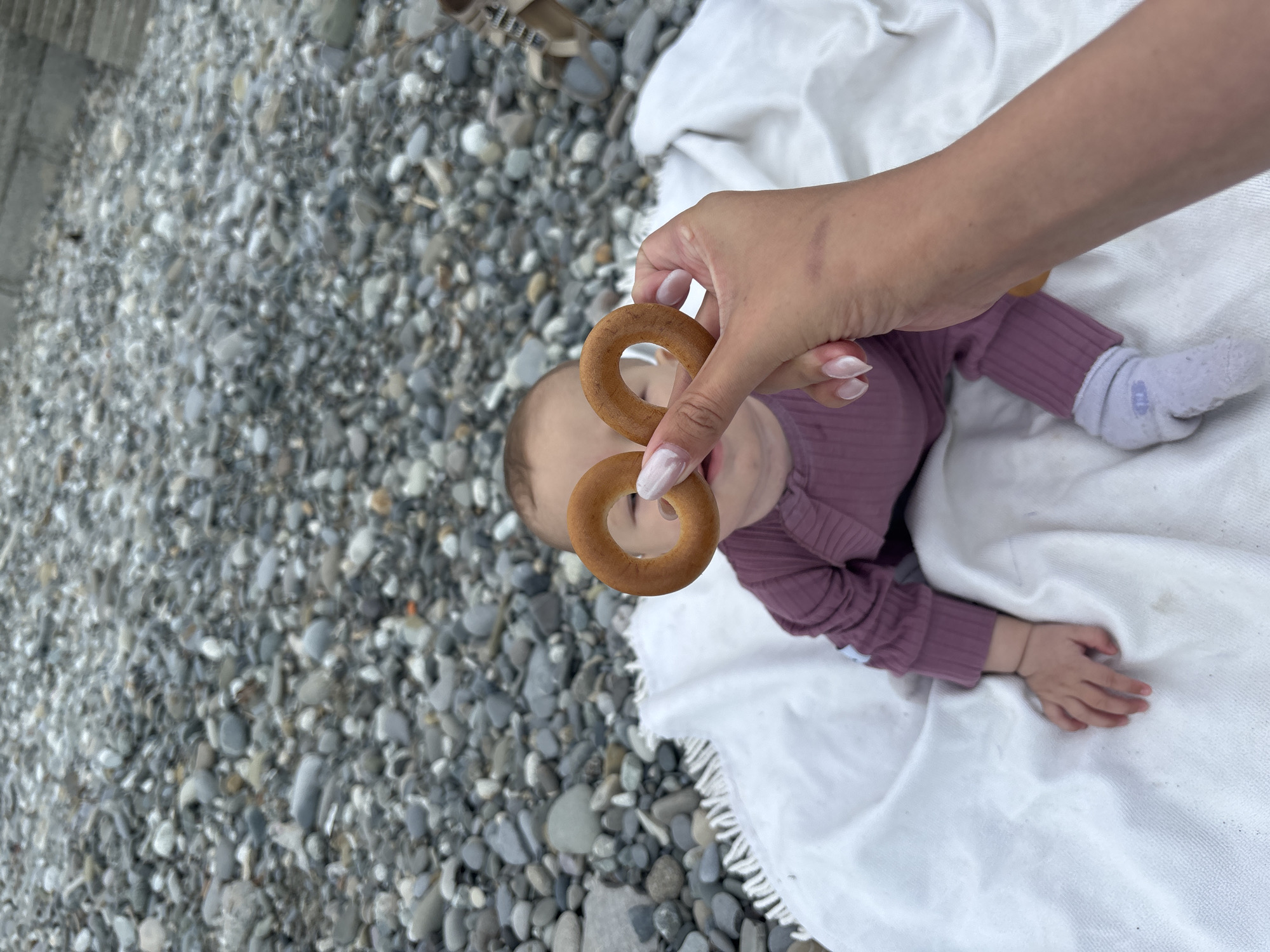 A top-down shot captures a baby lying on a white blanket with the lower half of the frame filled by a pebble beach. A woman's hand is holding two round, brown snacks, possibly bagels, in front of the baby's face, obscuring it. The baby is wearing a purple ribbed outfit and a white sock on one foot. The location is Sochi, Russia, evident from the characteristic pebble beaches found in the region.