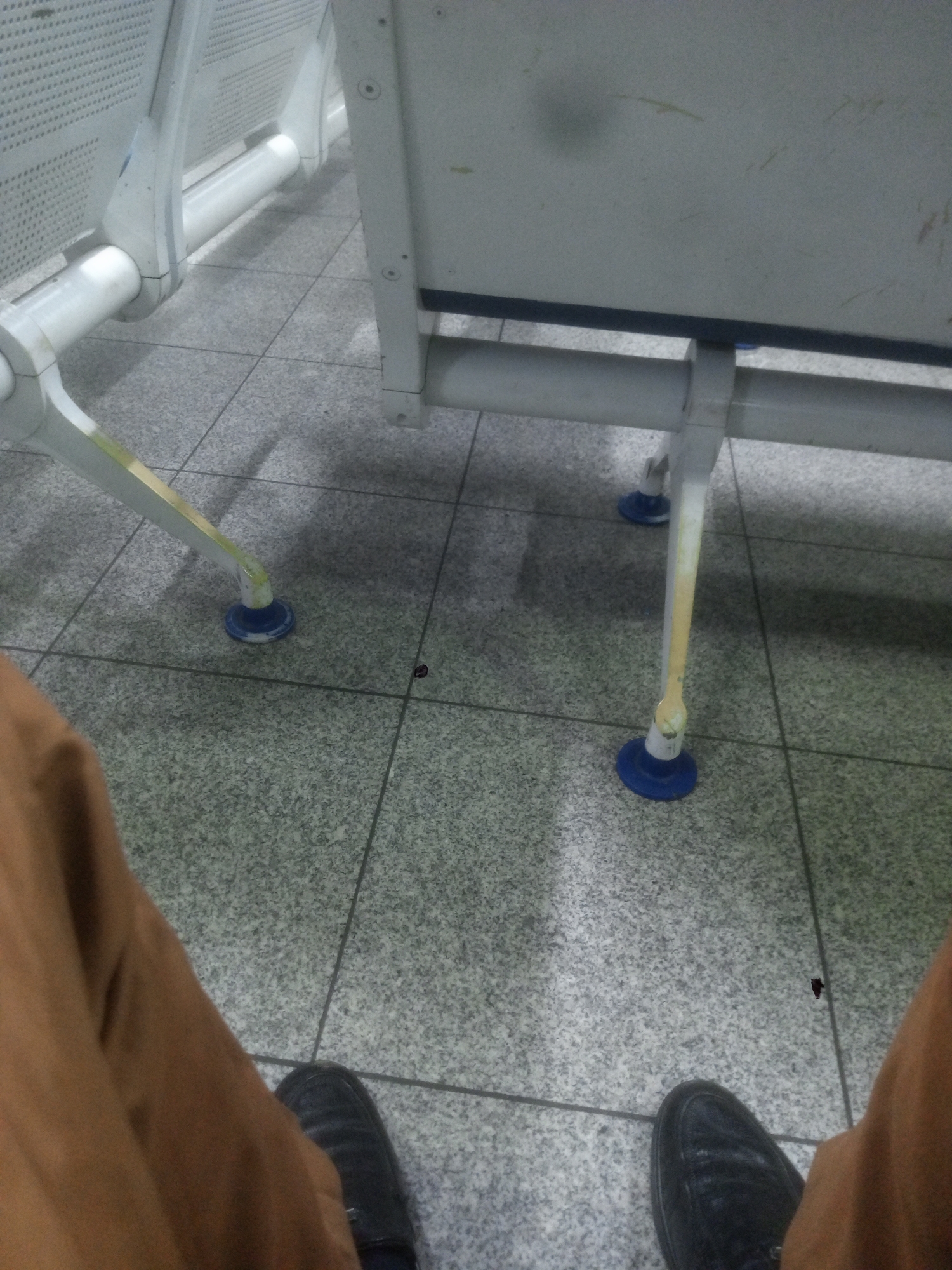 The image shows the lower portion of a person sitting on a public bench. The person is wearing brown pants and black leather shoes. The bench is white with blue leg caps, and sits on a gray tiled floor. Two small dark spots are also visible on the floor, near the person's legs. The location is not discernible from the image. The city of Gwaska, Nigeria, cannot be confirmed as the location.