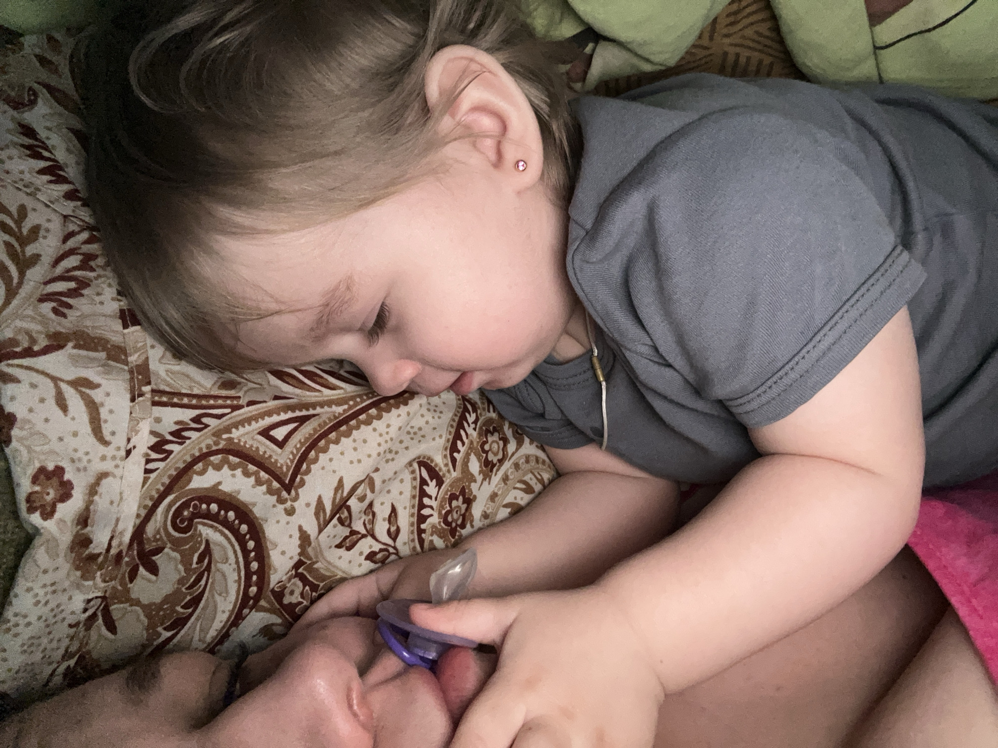 A young child with light brown hair and a small pink earring is lying on a patterned bedspread. They are wearing a gray t-shirt. The child's hands are holding a purple pacifier near the face of an adult. The adult's face is visible at the bottom of the frame. There is a green pillow visible in the upper right corner. The city of Chaikovsky, Russia is not discernible from the image.