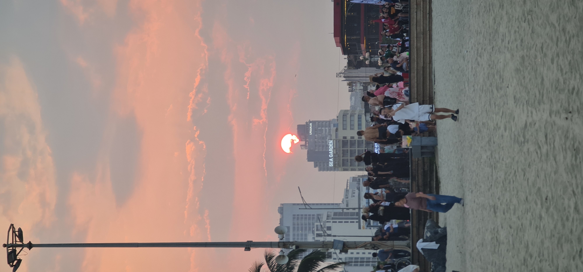 The image shows a beach in Đà Nẵng, Vietnam at sunset. The sun is setting behind several buildings, including one named "SEA GARDEN." A crowd of people is gathered along the beach. Some people are standing on a raised boardwalk next to the beach, while others are on the sand. A person in white shorts is walking on the beach. A palm tree is visible in the foreground and a tall light pole is on the left. The sky is filled with clouds, and the overall scene is bathed in warm, orange light.