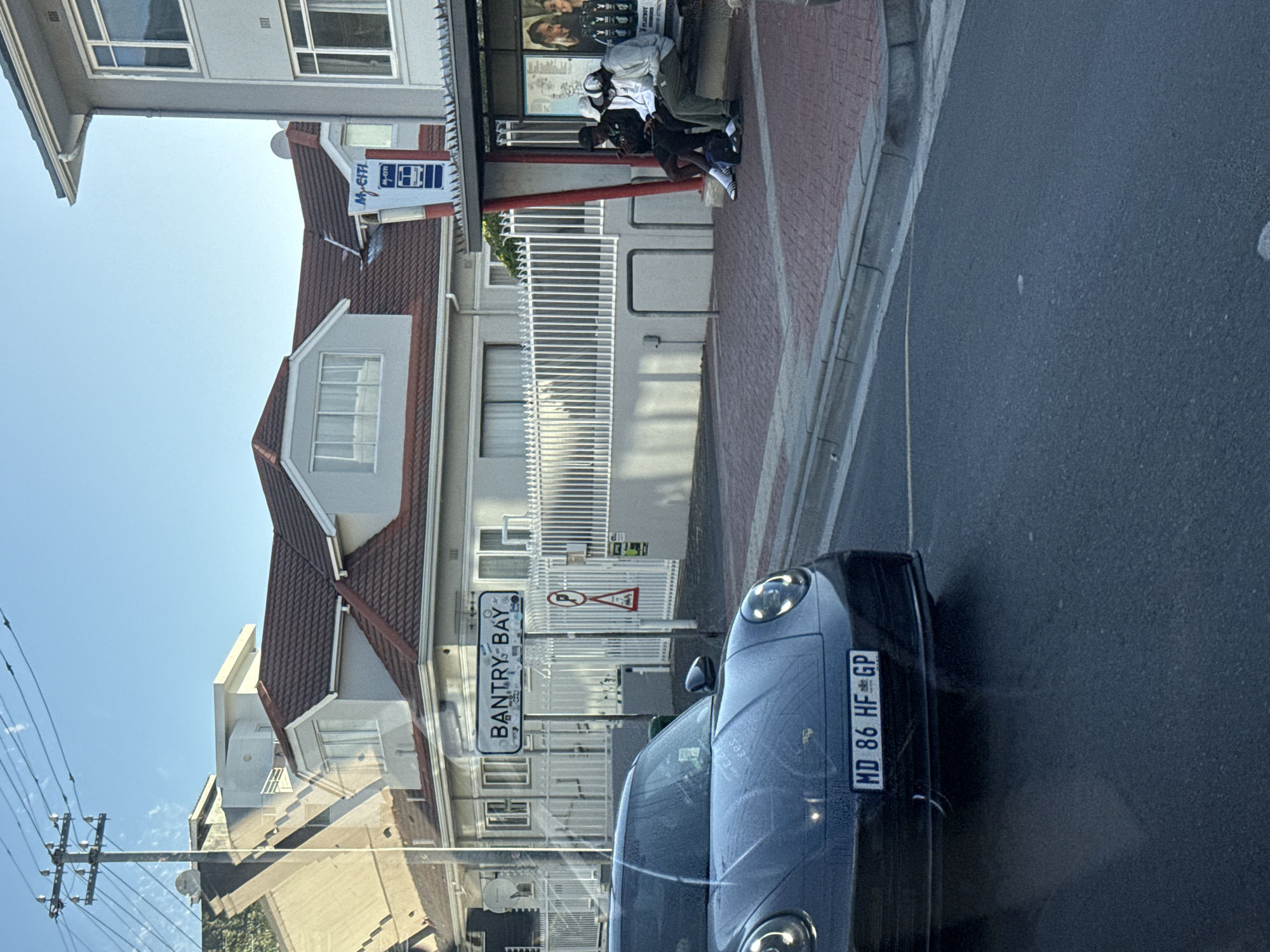 The image shows a street view in Bantry Bay, Cape Town, South Africa. A dark-colored car, with the license plate "MD 86 HF GP," is parked on the right side of the street. A street sign reading "Bantry-Bay" is visible. In the background, there is a building with a red-tiled roof and white walls. A bus stop with people sitting and standing is on the sidewalk. The sky is clear and blue. Overhead power lines are visible in the upper left corner.