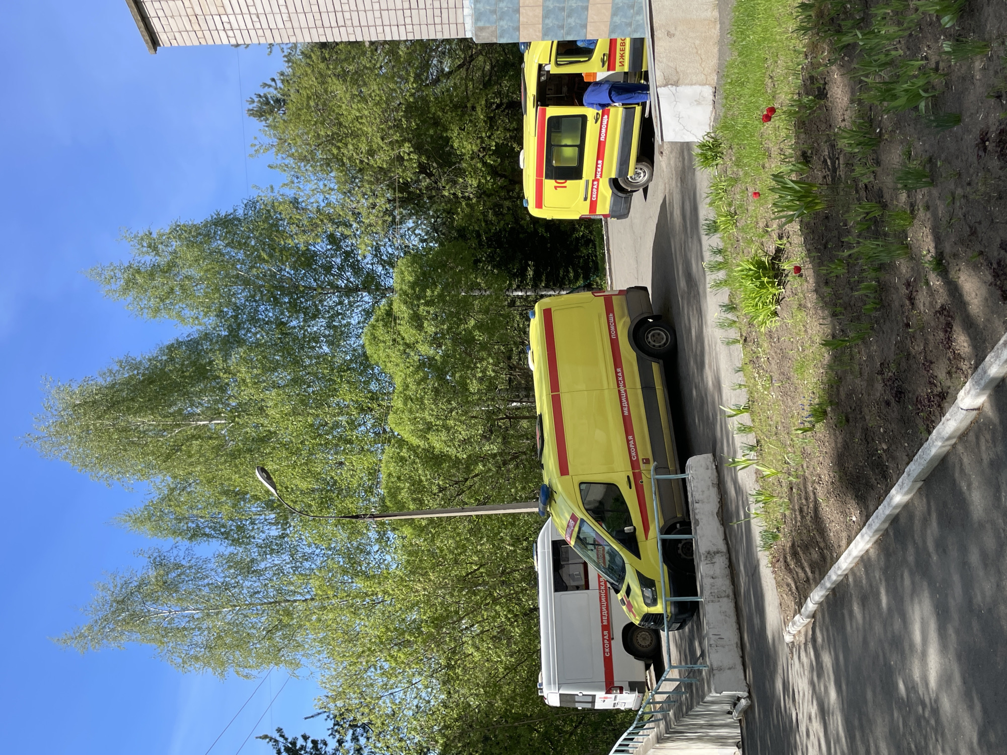 The image depicts a street scene in Izhevsk, Russia. Three ambulances are parked on the side of a road adjacent to a sidewalk and a low concrete barrier with metal railings. The ambulances are painted in white and yellow with red stripes and Cyrillic text that reads "Скорая Медицинская Помощь" (Emergency Medical Assistance) and "Ижевск" (Izhevsk). A person wearing blue clothing stands next to one of the ambulances. The environment includes trees, grass, and some red flowers. A street light is visible, as is part of a brick building and clear blue sky.