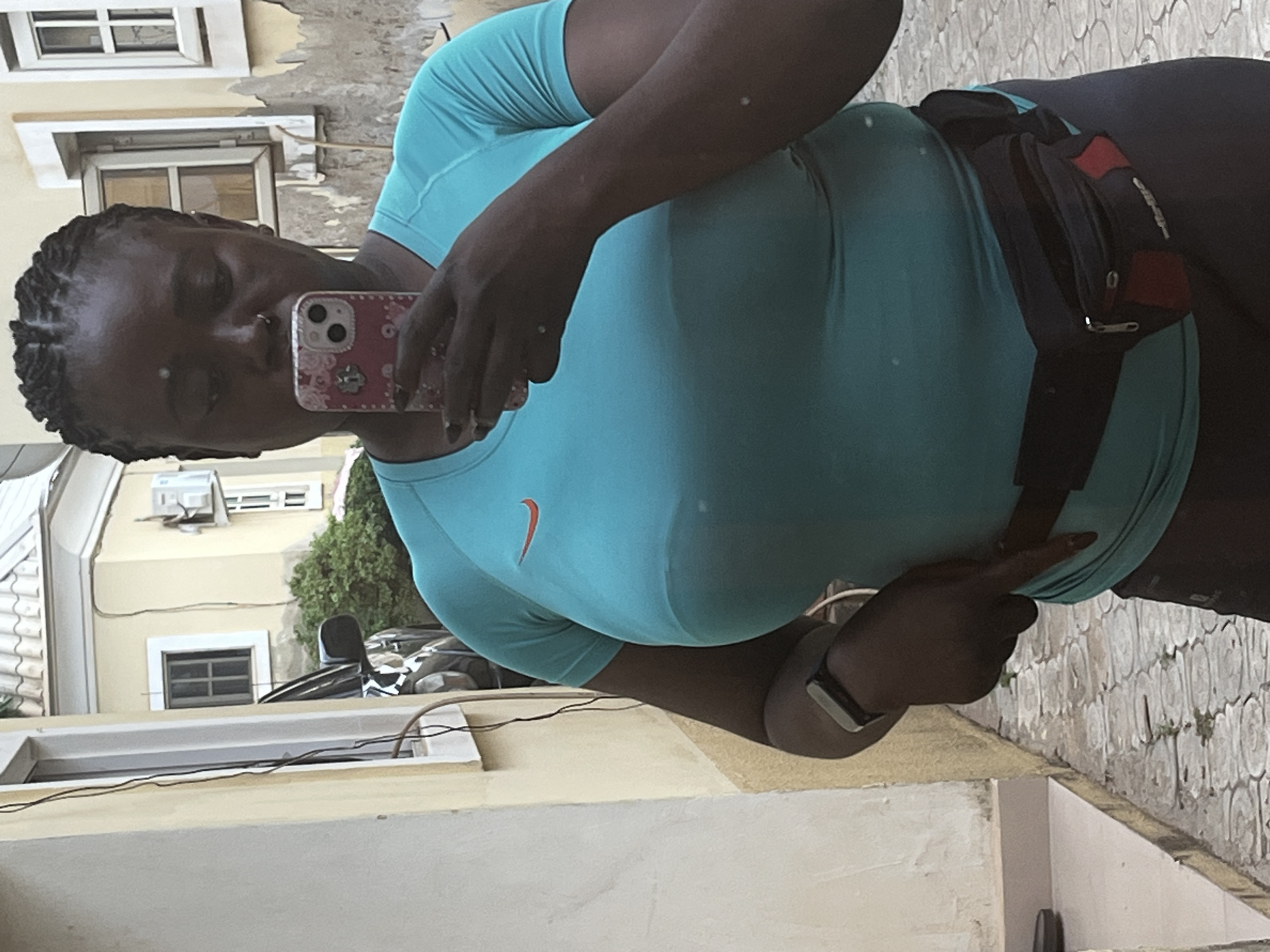 In an outdoor setting, a dark-skinned woman with braided hair is taking a selfie in front of a yellow building. She is wearing a teal athletic t-shirt with an orange Nike logo and dark leggings. A black fanny pack is worn around her waist. She has a watch on her left wrist and is holding a pink phone in her right hand. The background shows part of a building with windows and an air conditioning unit. The ground is paved with grey tiles. This scene is located in Abuja, Nigeria.