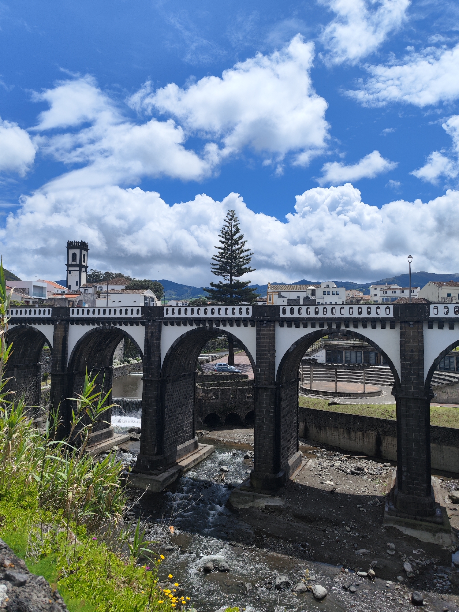This media file presents a vibrant, sunlit landscape centered on a historic stone bridge in Ribeira Grande, Portugal.
The main subject is a large, multi-arched bridge with dark stone foundations and pillars, contrasting with white painted sections and a distinctive black and white decorative railing along its top. The bridge spans a rocky riverbed where water flows, cascading over small weirs or waterfalls. Through the arches, parts of the riverbank, another smaller bridge, and an outdoor amphitheater-like structure are visible.
In the immediate foreground on the left, lush green vegetation, including tall reeds and small yellow and purple wildflowers, grows along a stone embankment. Beyond the bridge, a charming townscape emerges with white buildings, some featuring red-tiled roofs, backed by rolling green mountains. A prominent black and white church tower, with an ornate top, stands out on the left side of the town. A tall, slender conifer tree rises conspicuously behind the bridge in the center.
The sky is a brilliant blue, adorned with numerous large, fluffy white cumulus clouds, indicating clear and pleasant weather. The bright sunlight suggests it is daytime, likely late morning or early afternoon. No people are visible, and no specific activities are taking place, contributing to a serene and timeless atmosphere. Faint, indistinct text, possibly "SARA" or "SAPA," can be seen on a wall under one of the bridge arches.