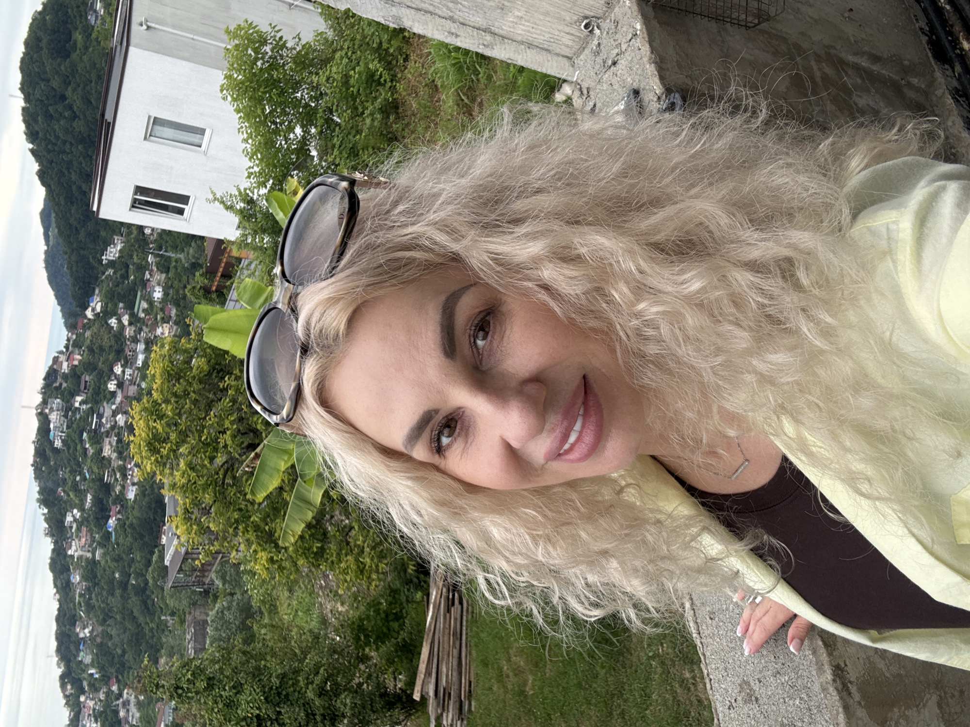 The media file is a selfie taken by a woman with long, blonde, curly hair, wearing sunglasses perched on her head. She is smiling at the camera, with her face in the foreground and right side of the frame. She is wearing a light yellow jacket over a dark brown top, and a delicate silver necklace. Her right hand is visible, resting on a grey, textured surface, and she is wearing a ring on her finger.
The background shows a scenic view of a hillside town, with many houses clustered on the slope. The houses appear to be of various colors and sizes, suggesting a residential area. Lush green trees and vegetation cover the hillside, with some large banana-like leaves visible in the middle ground. A white building with windows is situated higher up on the hill, partially obscured by trees. The sea or a large body of water is visible in the distance on the left. The scene suggests a warm, possibly humid day, with overcast or diffused lighting, making it difficult to pinpoint the exact time of day. The overall mood is relaxed and pleasant.
The location context provided is Baranovka, Russia, which aligns with the mountainous and coastal scenery depicted. There is no visible text in the image.