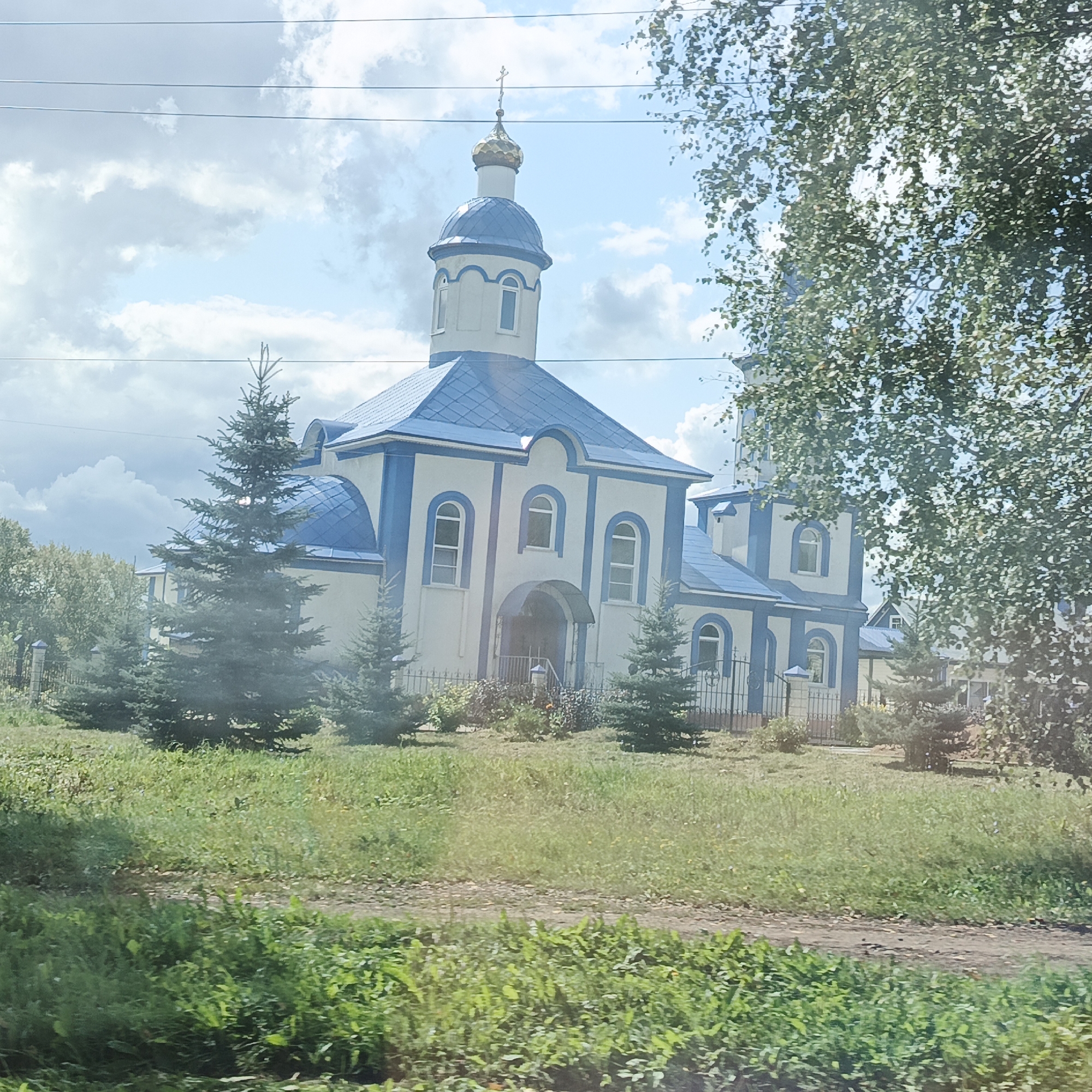 The image shows an Orthodox church with white walls and blue trim, featuring a central dome topped with a cross and several smaller domes. The church has arched windows and a prominent entrance. It is surrounded by green grass, several evergreen trees, and foliage. In the background, there are more trees and buildings, suggesting a rural or suburban setting. The sky is partly cloudy with a bright, sunny appearance. The scene appears to be captured on a clear day. Based on available information, this church is likely the Church of the Resurrection of Christ located in Chishmy, Russia.