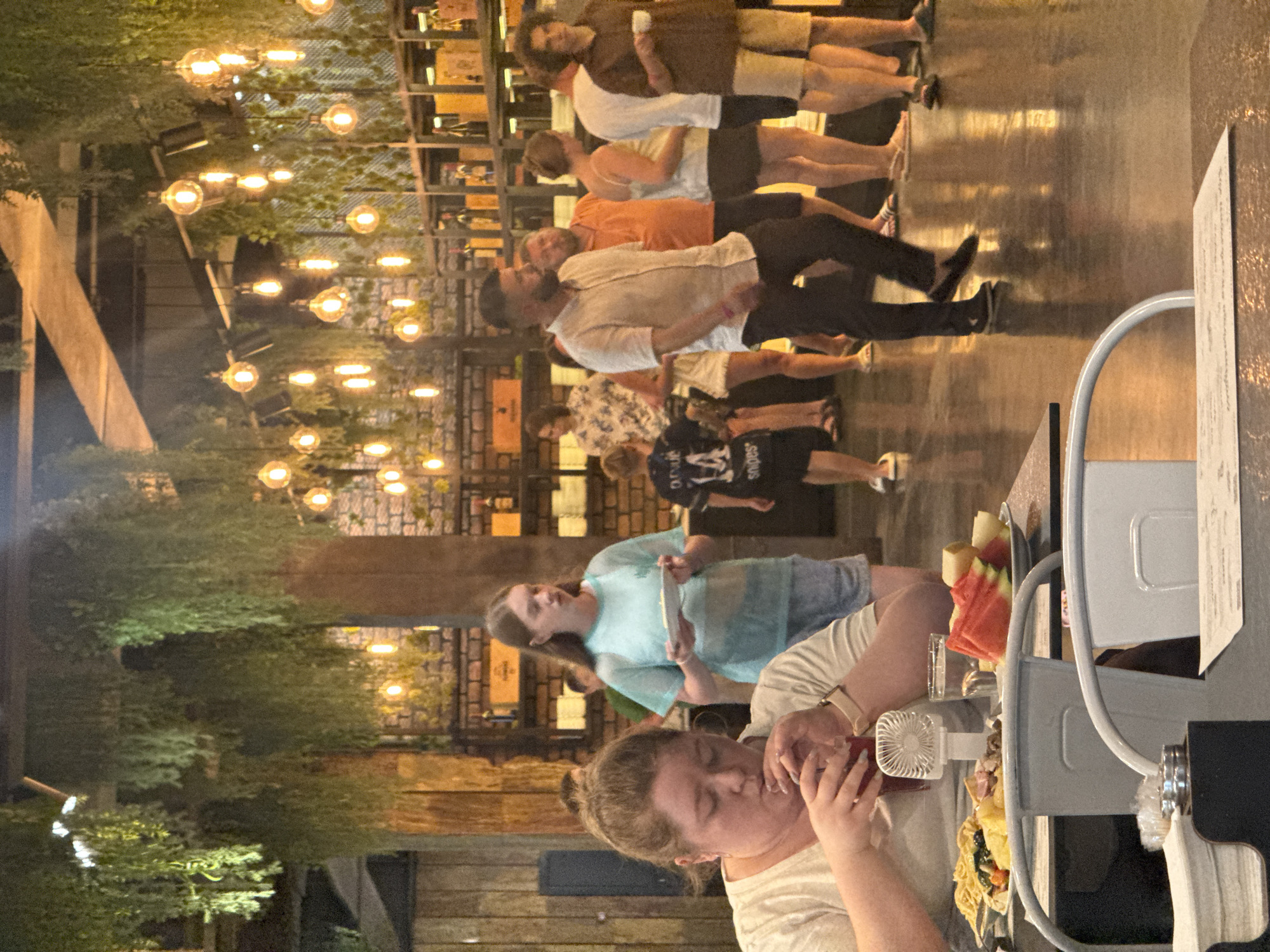 In a dimly lit restaurant, likely in the city of Yalta, Ukraine, patrons are gathered. In the foreground, a woman with light brown hair is seated and drinking from a glass. In front of her is a table with a plate of food, including pasta and fruit, and a small portable fan. Behind her, a woman in a light blue shirt holds a white plate. Further back, a group of people are standing, some in conversation. The restaurant features exposed brick walls, hanging plants, and numerous decorative light bulbs suspended from the ceiling. The floor appears to be polished concrete, reflecting the ambient light.