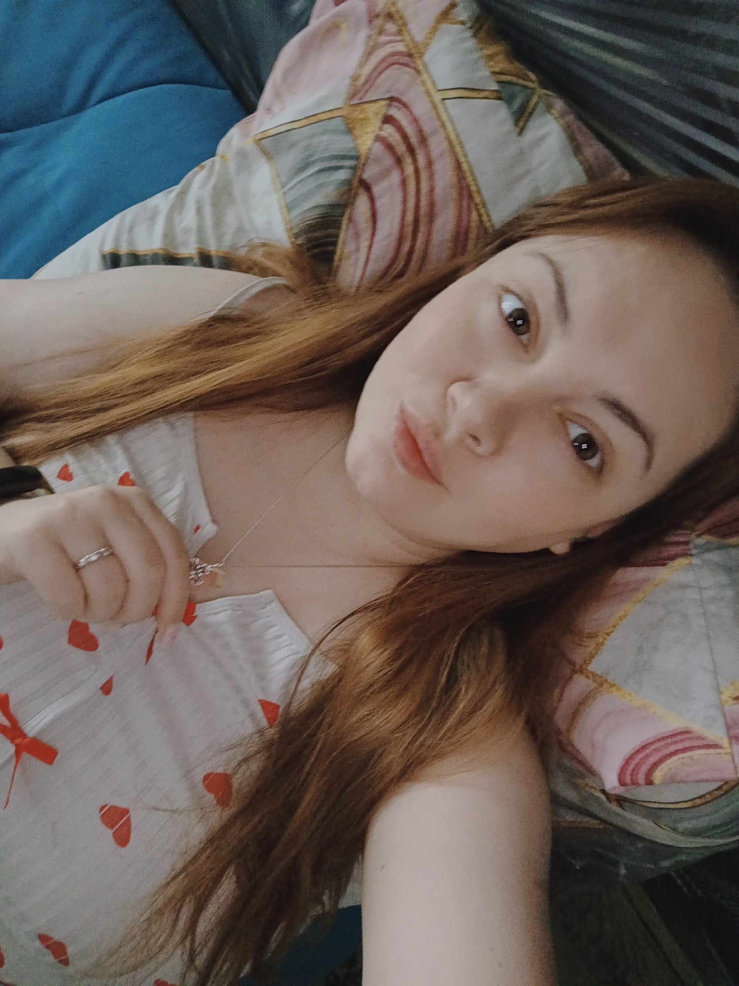 A woman lies on a bed. She has brown hair and wears a white top with red hearts. She holds a necklace and wears a ring on her left ring finger. The bed has a blue pillow and a patterned pillow. The location is presumed to be in Chaikovsky, Russia.