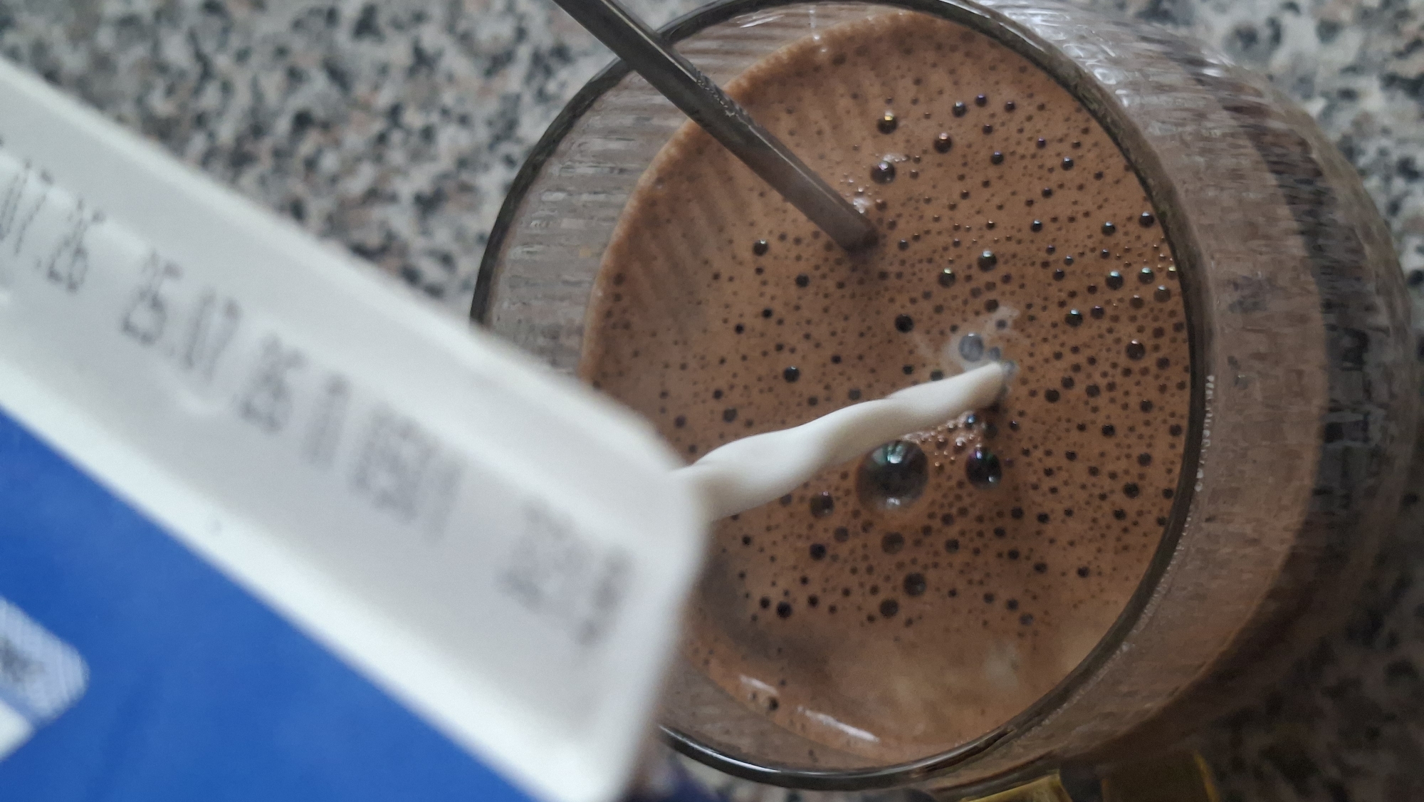 A carton of milk is being poured into a glass of chocolate drink. The drink is a rich brown color and has a foamy top with many small bubbles. A silver spoon is submerged in the drink, partially obscured by the stream of milk. The carton is white with blue and black text. The background is a speckled gray surface, possibly a countertop. The image is a close-up shot, with the focus on the pouring action and the drink.