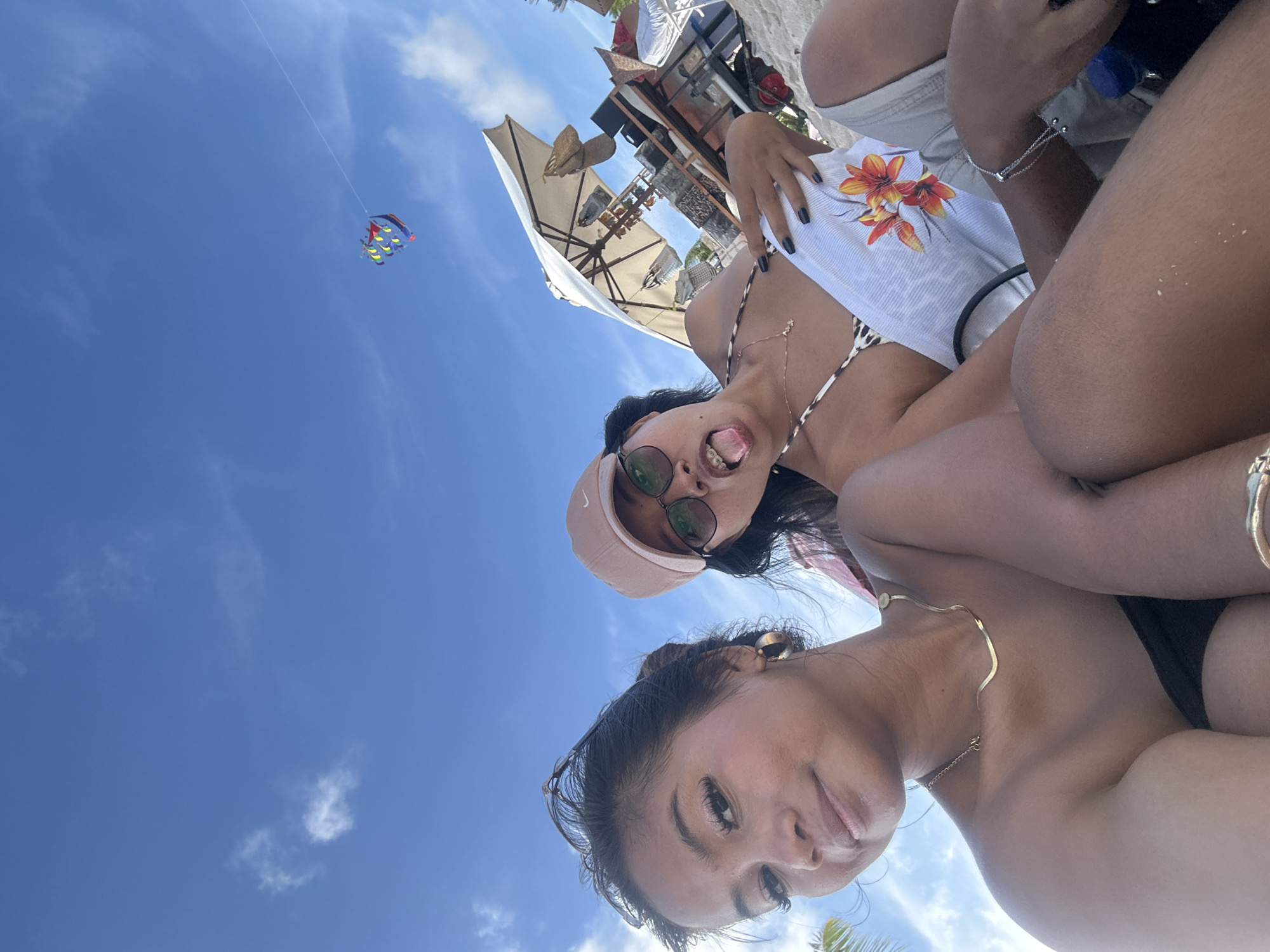 The photograph appears to be taken from a low angle on a beach in Kuta Selatan, Indonesia, based on the presence of beach umbrellas, sky, and sand. Two women are prominently featured in the foreground. One woman is wearing a pink cap and sunglasses, sticking her tongue out playfully. The other woman has her hair pulled back and wears a gold necklace. Both women seem to be sunbathing. The background features a building or structure with white umbrellas. Above the two women, in the sky, is an object which is possibly a kite being flown. The environment suggests a sunny day on a tropical beach.