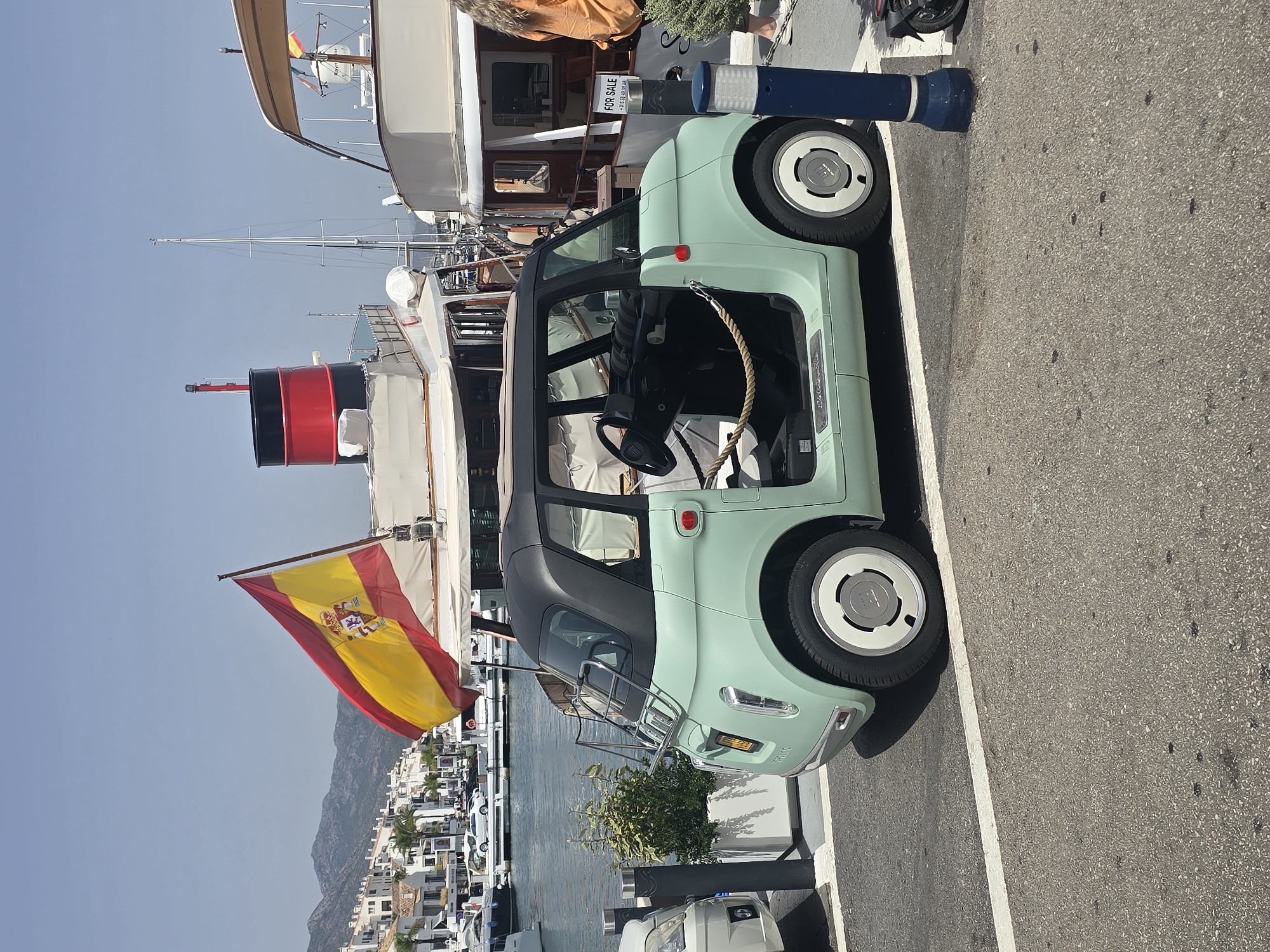 This is a full-shot photograph of a light green, two-door compact electric vehicle parked on an asphalt surface next to a marina in Marbella, Spain. The car is a "NISSA" model, and it has a black roof and black interior. The driver's door is open, revealing the interior and a rope barrier. A Spanish flag is prominently displayed on a pole behind the car, and it is unfurled in the wind. Several yachts and boats are docked in the marina, and in the background, white buildings with tiled roofs and mountains are visible under a clear blue sky. The scene is brightly lit by sunlight, suggesting it is daytime. A sign on one of the yachts reads "FOR SALE".