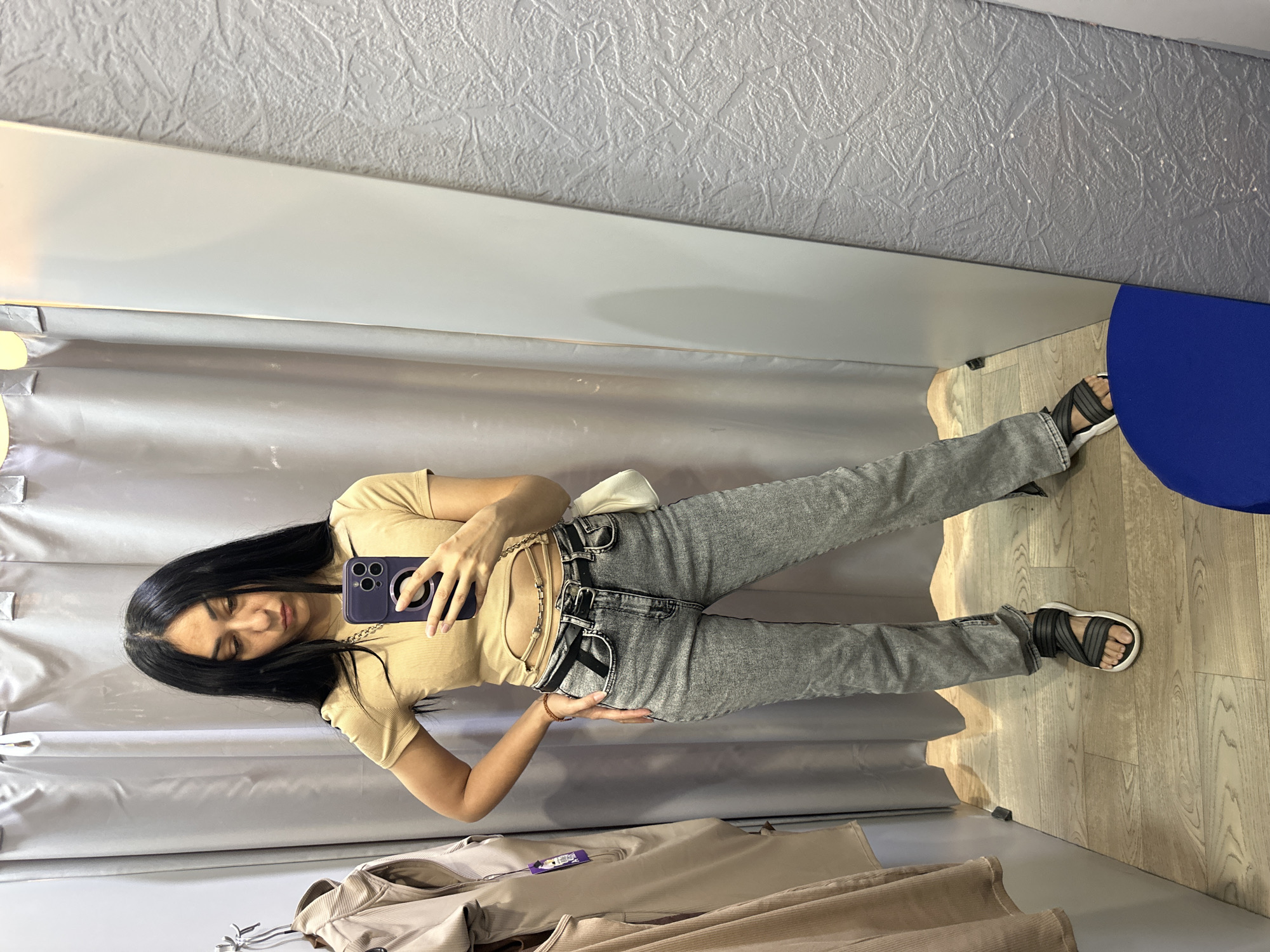 A young woman with long, dark hair is posing for a selfie in a fitting room. She is wearing a fitted, light brown, short-sleeved t-shirt, grey jeans, and black sandals with white soles. A small, white pouch is clipped to her belt. She holds a dark purple smartphone in her right hand, taking the photo in a large mirror that reflects the fitting room. The fitting room has textured grey walls and a light wood-grain floor. Behind her, there are light grey curtains. To the right of the frame, a large, bright blue circular object is visible, possibly a piece of furniture or display. The overall lighting suggests an indoor setting, likely during the day. The woman appears to be looking down at her phone with a slight smile, perhaps assessing her outfit. Another piece of clothing, a light brown jacket or coat, is draped on a rack in the foreground, with a purple tag attached.