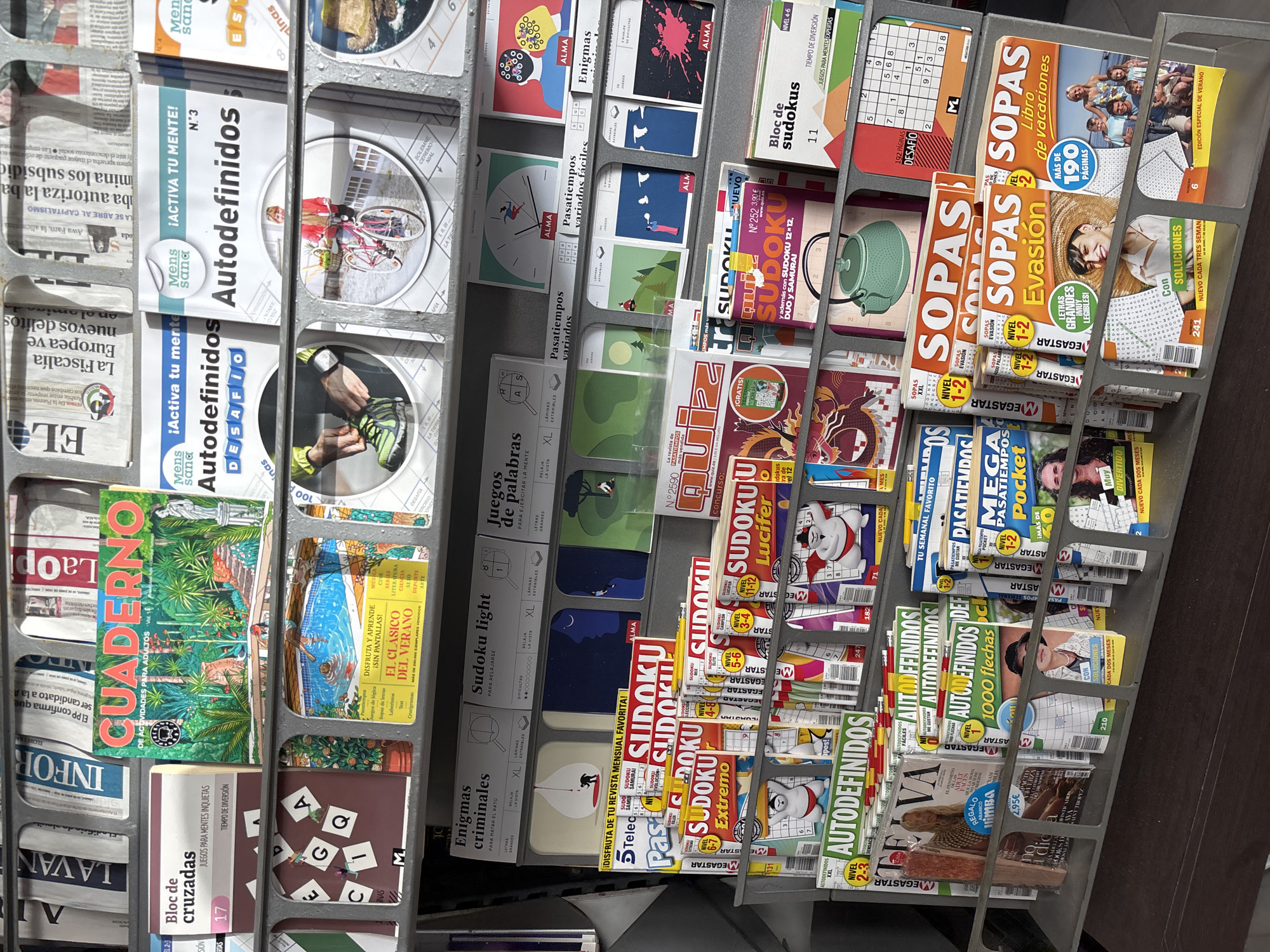This image depicts a display rack filled with magazines and puzzles in a retail setting, likely a newsstand or magazine store. The products are predominantly puzzle books and magazines focused on logic games and mental challenges, such as Sudoku and Autodefinidos (crosswords). Many of the publications are in Spanish, indicated by titles like "CUADERNO DE ACTIVIDADES PARA ADULTOS," "Juegos de Palabras," and "El Clásico del Verano."
The display is organized in horizontal tiers, with each tier holding multiple items. Some publications are stacked horizontally, while others are displayed vertically, revealing their covers and spines. The background consists of other magazines and newspapers, some with headlines visible, suggesting a broad selection of reading material. The lighting is bright and even, illuminating the products clearly.
The lower section of the display shows more magazines, including titles like "SOPAS Evasión" and "SOPAS Libro de vacaciones." These also feature puzzles and activities. The overall impression is a well-stocked retail display aimed at customers seeking entertainment and mental stimulation through word and number games. The scene is clean and orderly. No specific geographic markers like street signs or distinctive architecture are visible within the frame, therefore, its specific location in Orihuela, Spain cannot be determined from the image itself.