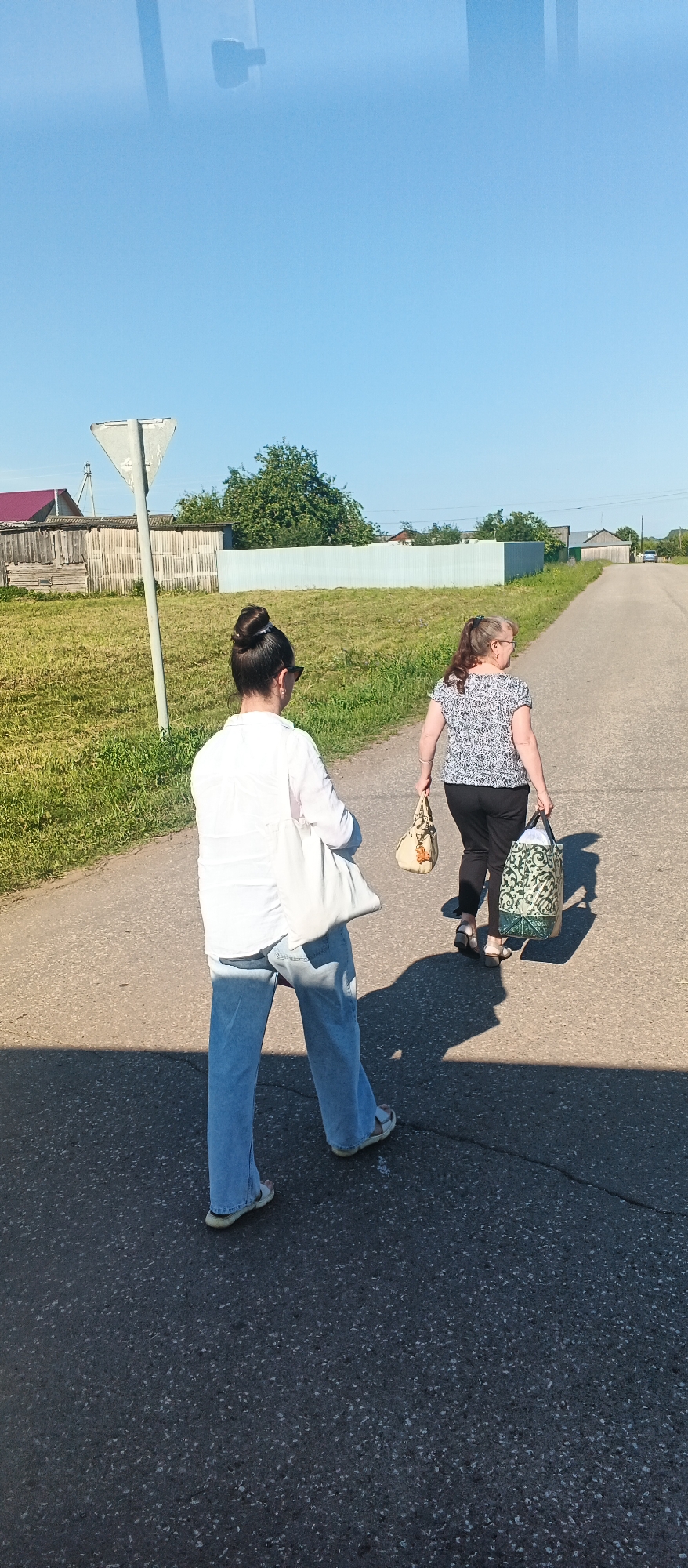 Two women are walking on a paved road in what appears to be a rural or semi-rural area, possibly in the village of Kiyasovo, Russia, based on the context. The woman in the foreground is facing away from the camera, wearing a white long-sleeved shirt, light blue jeans, and sandals. She has her dark hair pulled back in a bun and is carrying a large, light-colored tote bag over her left shoulder. The second woman is ahead of her, also facing away, wearing a black and white patterned short-sleeved shirt, black pants, and sandals. She is carrying two bags: a beige purse in her left hand and a larger, patterned tote bag in her right hand. The road is a two-lane asphalt surface with a visible crack. To the left of the road is a grassy area with a triangular road sign and a wooden fence in the background, behind which are some houses with red roofs. To the right of the road, the landscape is also grassy, leading to houses and trees in the distance. The sky is clear and blue, indicating a sunny day. The sun is casting long shadows of the women and the road sign onto the asphalt.