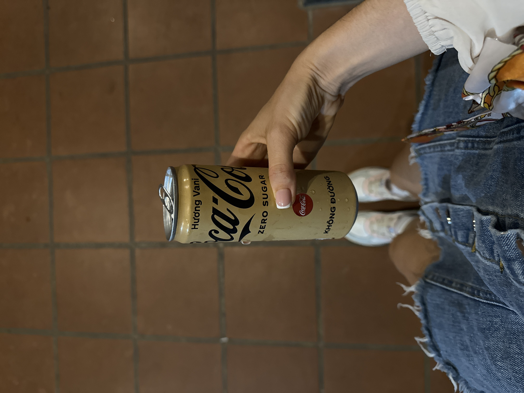 A person holds a can of Coca-Cola Zero Sugar Hương Vani in their right hand. They stand on a brick-tiled surface. The person is wearing denim shorts and white sneakers. The location is likely Mũi Né, Vietnam, given the Vietnamese text on the can.