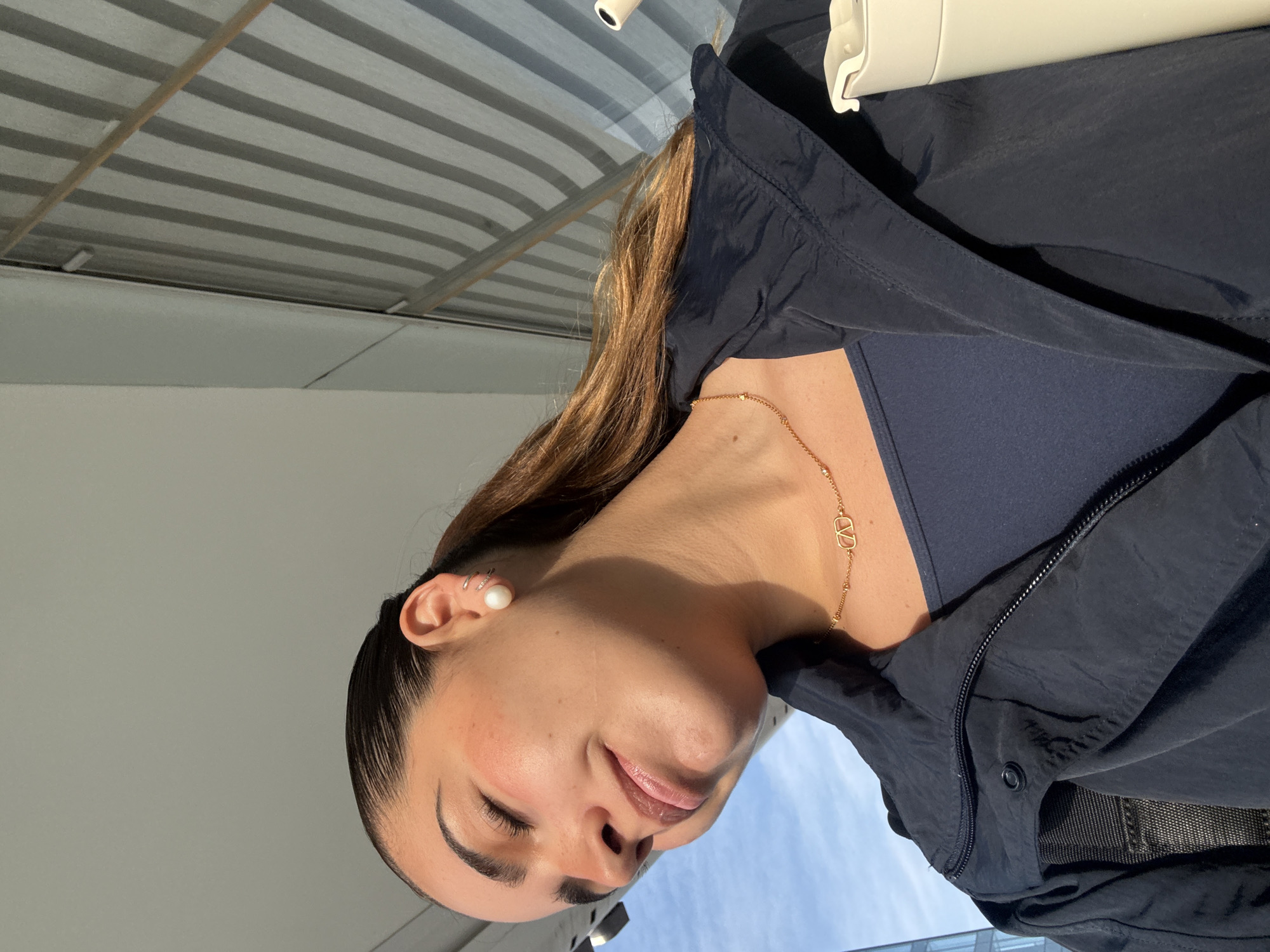 This media file presents a low-angle portrait of a young woman, likely taken outdoors or in a well-lit semi-enclosed environment in Puebla, Mexico, during daylight. The woman, with a light to medium complexion and long brown hair pulled back, is depicted from the chest up, looking downwards with her eyes closed, conveying a peaceful and serene emotion.
She is dressed in a dark blue or black lightweight jacket, possibly a windbreaker, worn over a matching undershirt. Her accessories include a delicate gold chain necklace with an interlocking "CD" or similar logo-style pendant, likely from Dior. On her visible left ear, she wears a large pearl stud earring in the lobe and multiple small, shiny hoop or stud earrings along the cartilage. Subtle makeup, including blush and lip gloss, enhances her features.
The background above her head reveals a striped architectural element, possibly a corrugated metal ceiling or awning with alternating grey and white horizontal patterns. Below her, a section of clear blue sky is visible, suggesting good weather, along with a blurred glimpse of a building or structure in the distance. A white, unidentified object, potentially a bottle or part of a hanger, is partially seen in the upper right. The bright, natural light illuminates her face, creating soft shadows.