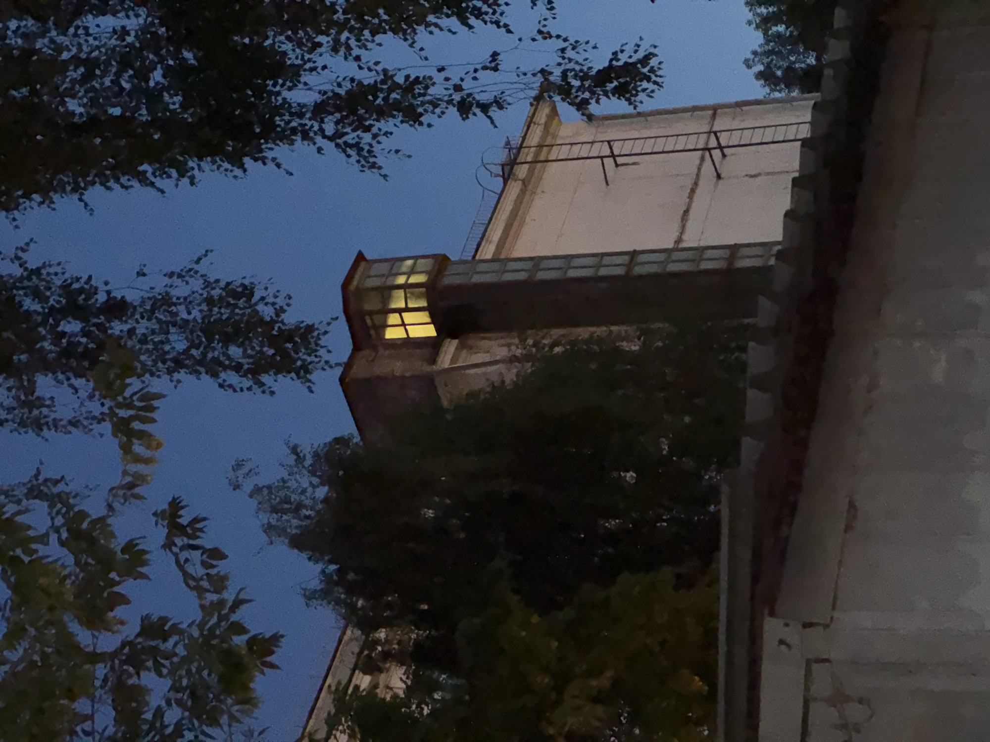 The image depicts the side of a multi-story building in Moscow, Russia, at dusk. The sky is a deep blue, suggesting the transition from day to night. In the foreground, a dark, textured roofline is visible, possibly belonging to a lower structure.
The main subject is the building itself, which appears to be constructed of concrete or similar material, painted a pale, off-white color. A prominent feature is a tall, narrow tower-like extension on the side of the building. This extension is primarily glass-paned, with vertical divisions. At the very top of this tower, a small, enclosed room or alcove is illuminated with a warm, yellow light, indicating occupancy or an interior light source. Below this illuminated section, the tower has a series of vertical, rectangular windows that are dark, suggesting they are either unlit or the interior is not visible.
A metal ladder is attached to the side of the building, extending upwards towards the roofline of the main structure. The ladder is black and appears to be a fire escape or maintenance access.
The building is partially obscured by dark, leafy trees and branches in the foreground and to the sides. These trees are silhouetted against the sky and the building, adding depth and a sense of being in a natural or semi-urban environment. The foliage appears to be deciduous, suggesting it could be autumn.
The overall impression is a quiet, perhaps slightly melancholic, urban scene at the end of the day. There are no people visible in the image.