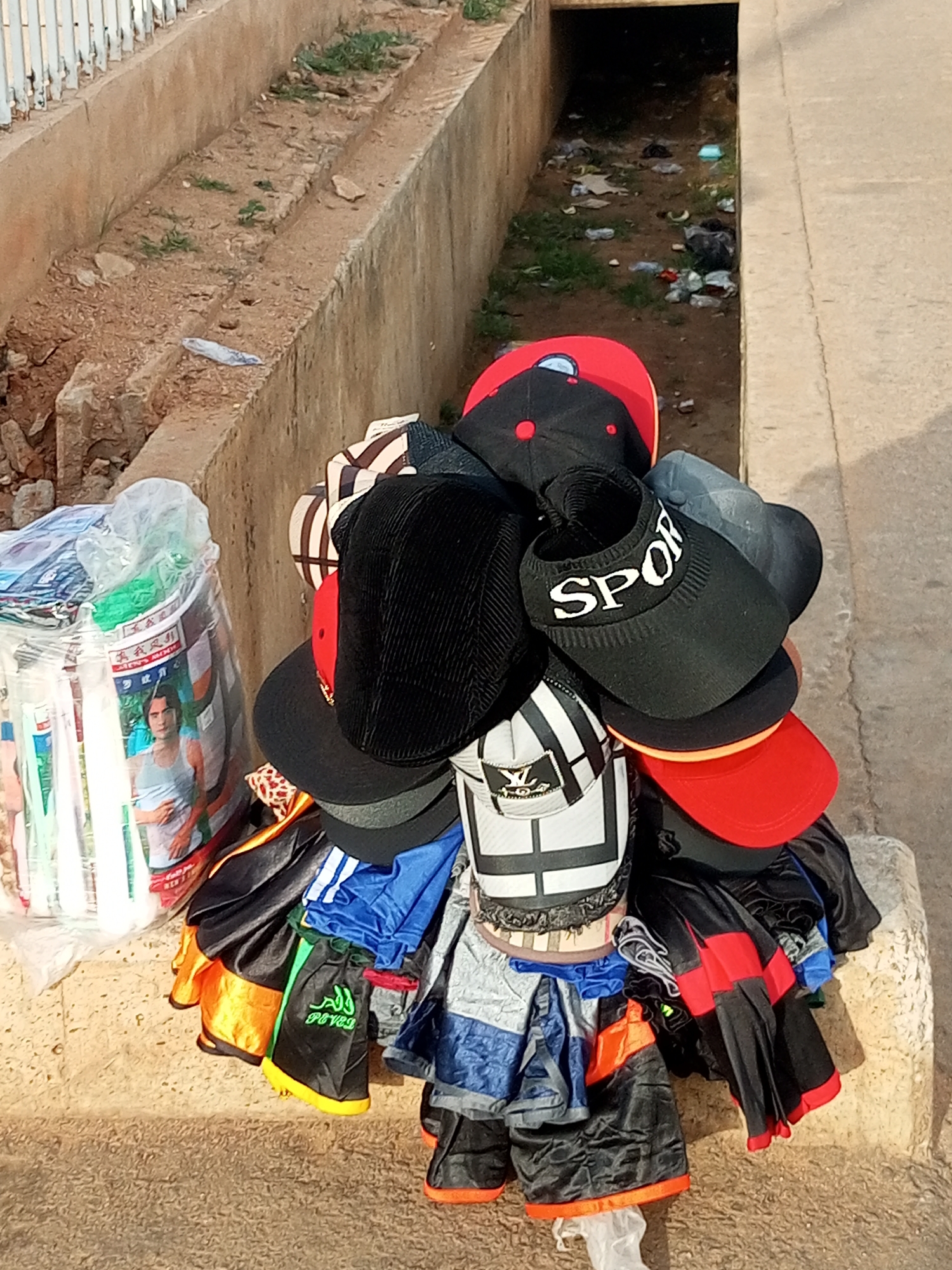 A vendor displays a variety of hats and shorts for sale. The hats, predominantly baseball caps and visors, are stacked in a large cluster. Some are plain black or red, while others feature stripes or the word "SPORT" in white lettering. Below the hats, several pairs of shorts are bundled together. They are in various colors, including black, blue, orange, and green, with some featuring white stripes or visible text, like the word "POVELL" on a green pair.
To the left of the hat display is a large, clear plastic bag containing more items, possibly clothing or accessories, with a picture of a young man in a white tank top visible on the packaging.
The scene is outdoors, with a concrete structure and a ditch filled with debris in the background. The ground is dirt and gravel. The lighting suggests it is daytime. The overall impression is of a street market or roadside stall.